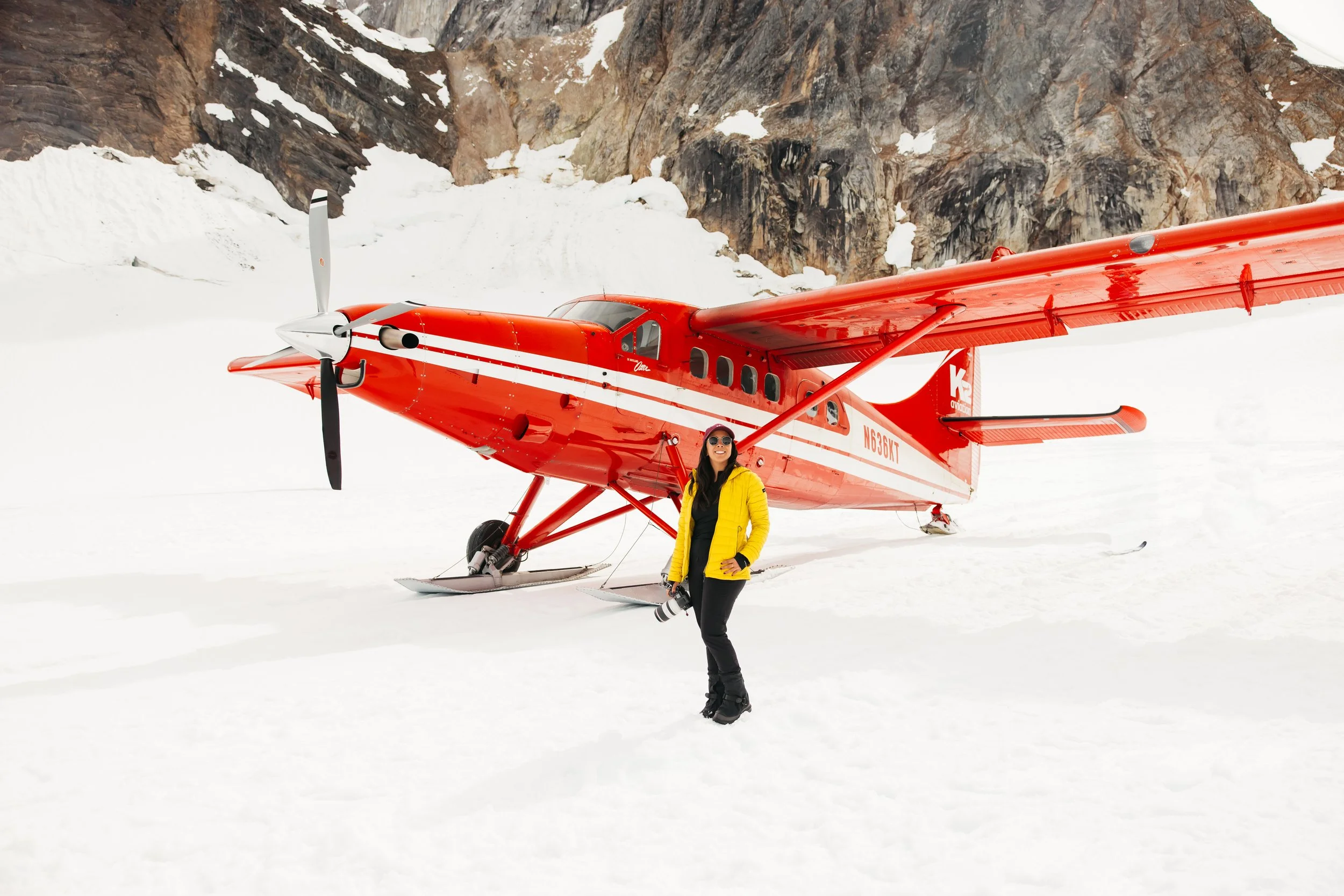 Traveler standing on a glacier beside a red K2 Aviation ski plane during the Denali Flyer tour in Alaska.