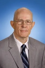 An elderly woman with short gray hair and glasses standing in front of a brick wall, wearing a red shirt and a music note decorated vest.