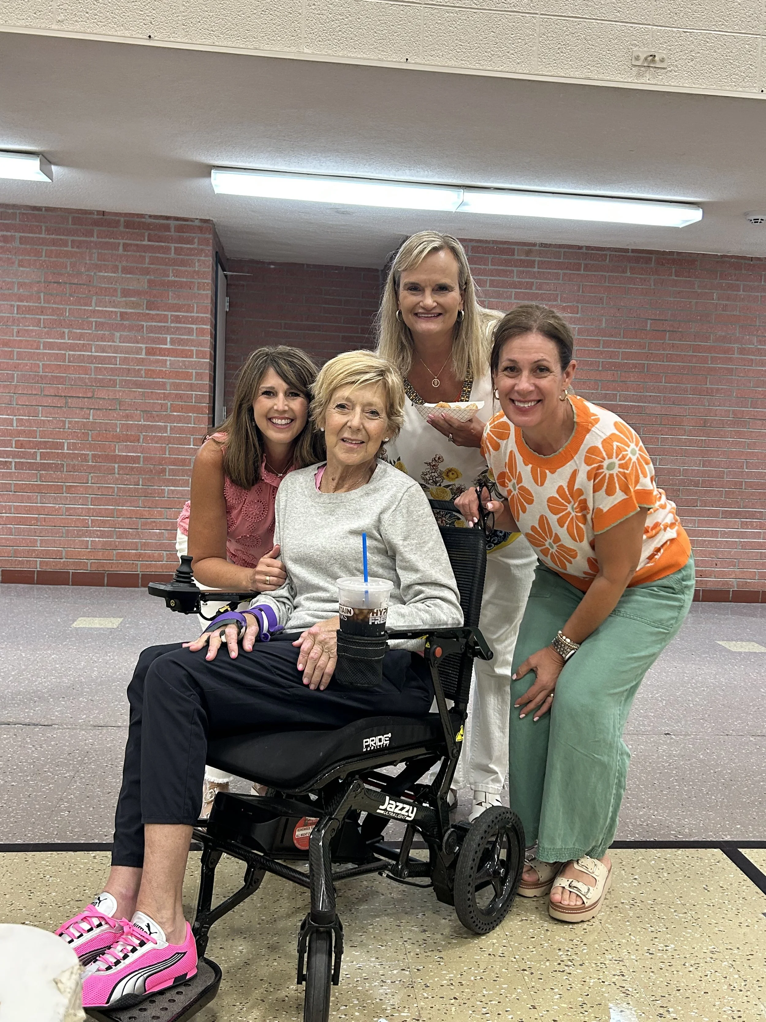 Group of five women, one in a wheelchair and one holding a bowl, smiling indoors against brick wall, casual attire.