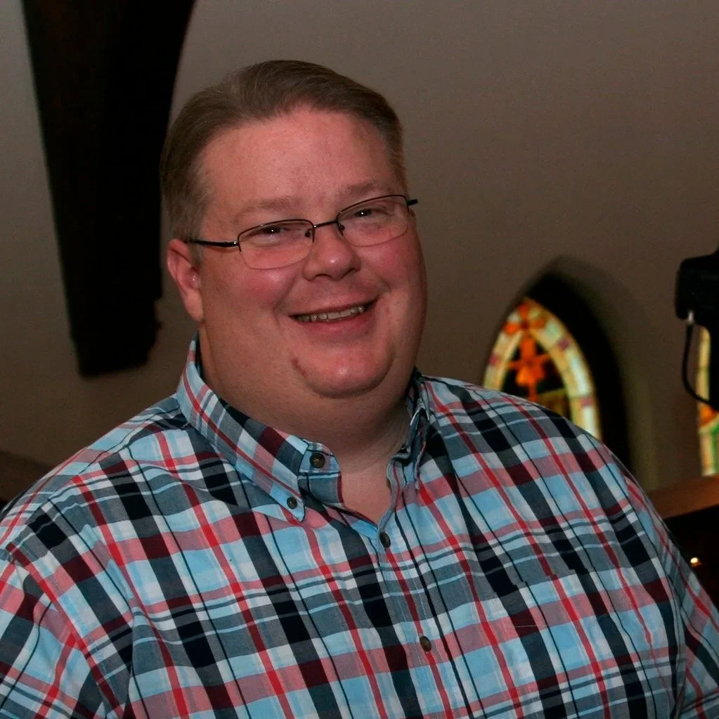A man smiling in front of a sound mixing console inside a church with stained glass windows.