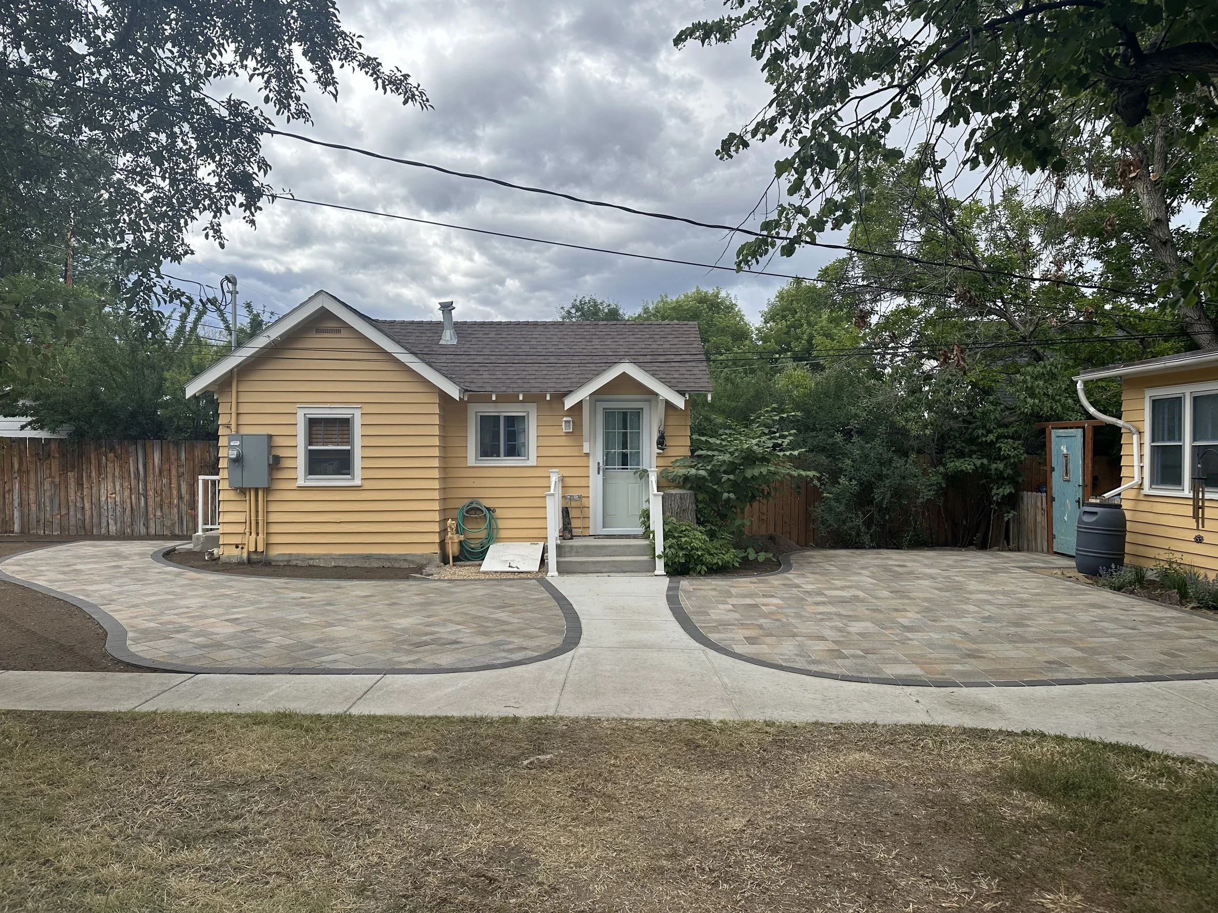 Front view of a small yellow house with a new paved driveway, stairs, and landscaping, under a cloudy sky.