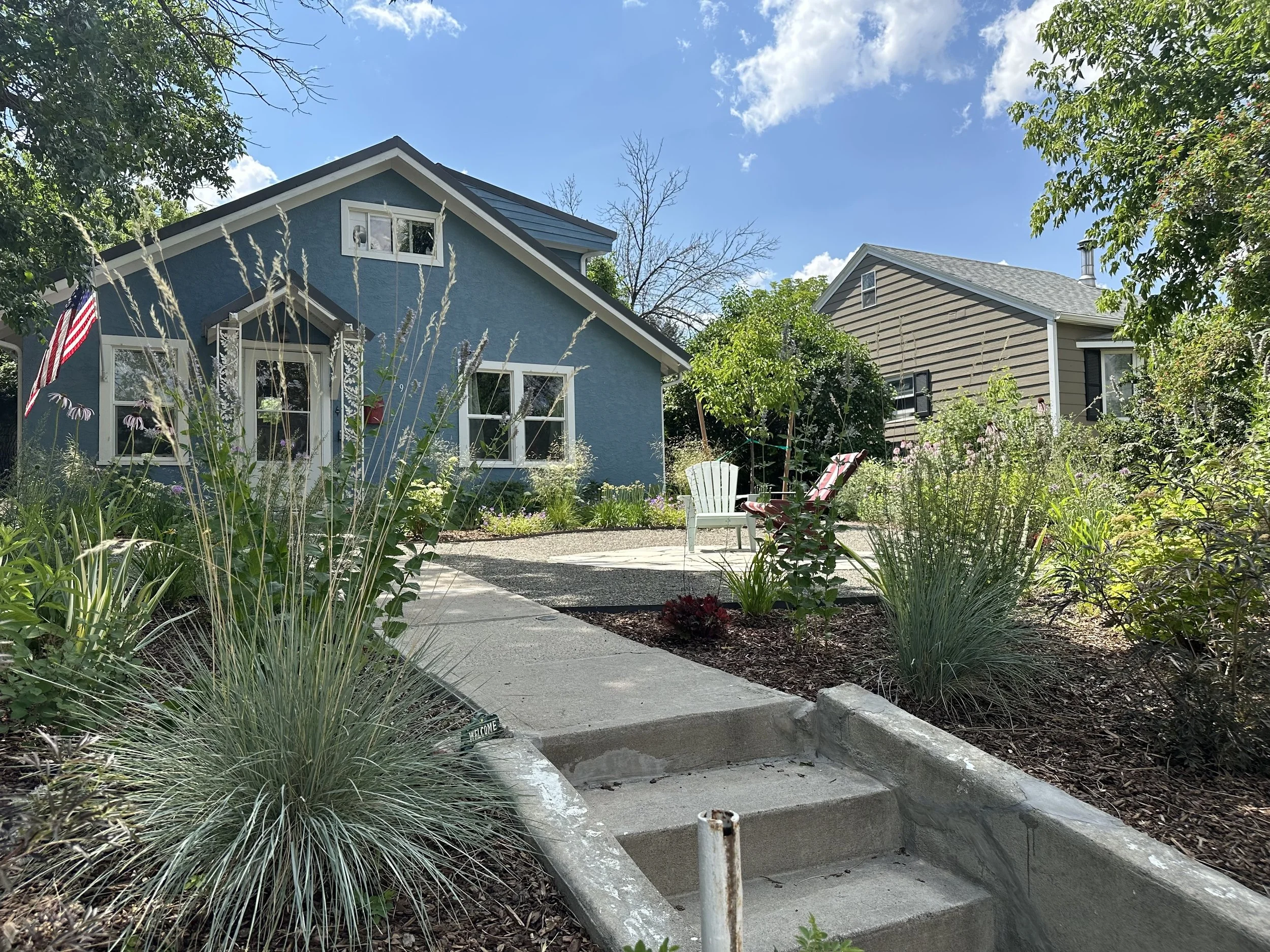 A suburban backyard with a blue house, a gravel patio, and lush greenery, including plants, trees, and garden chairs under a blue sky with some clouds.
