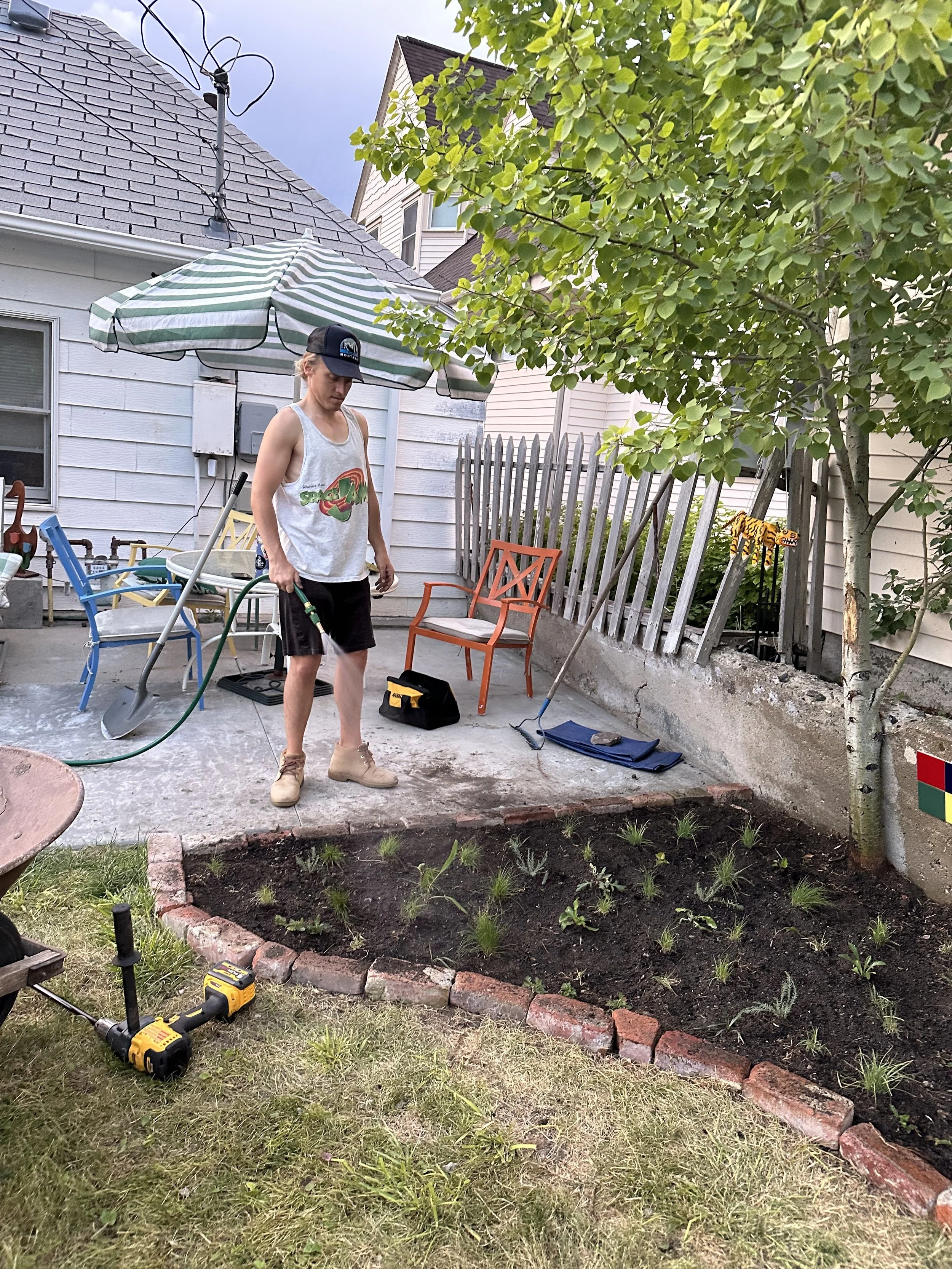 A young man watering a flower bed in a backyard with a garden hose, surrounded by outdoor furniture and tools.