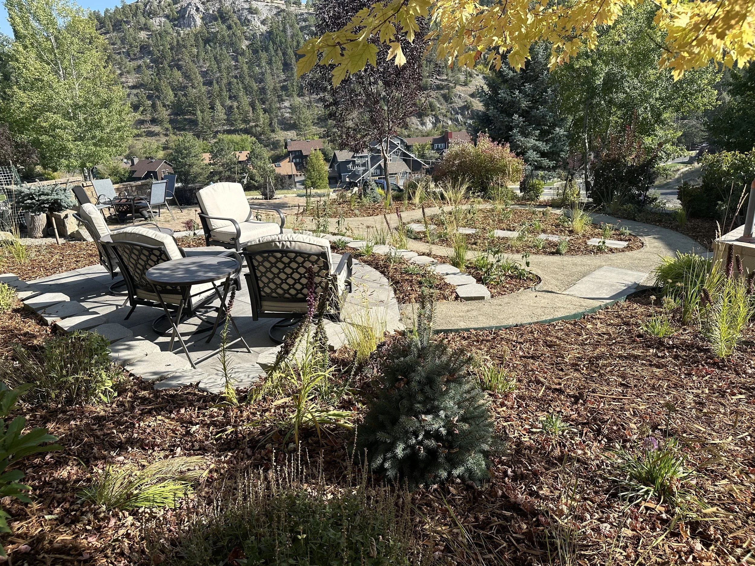 A landscaped backyard with a circular patio of chairs and table, surrounded by mulch and plants, with a curved pathway and houses on a hillside in the background.