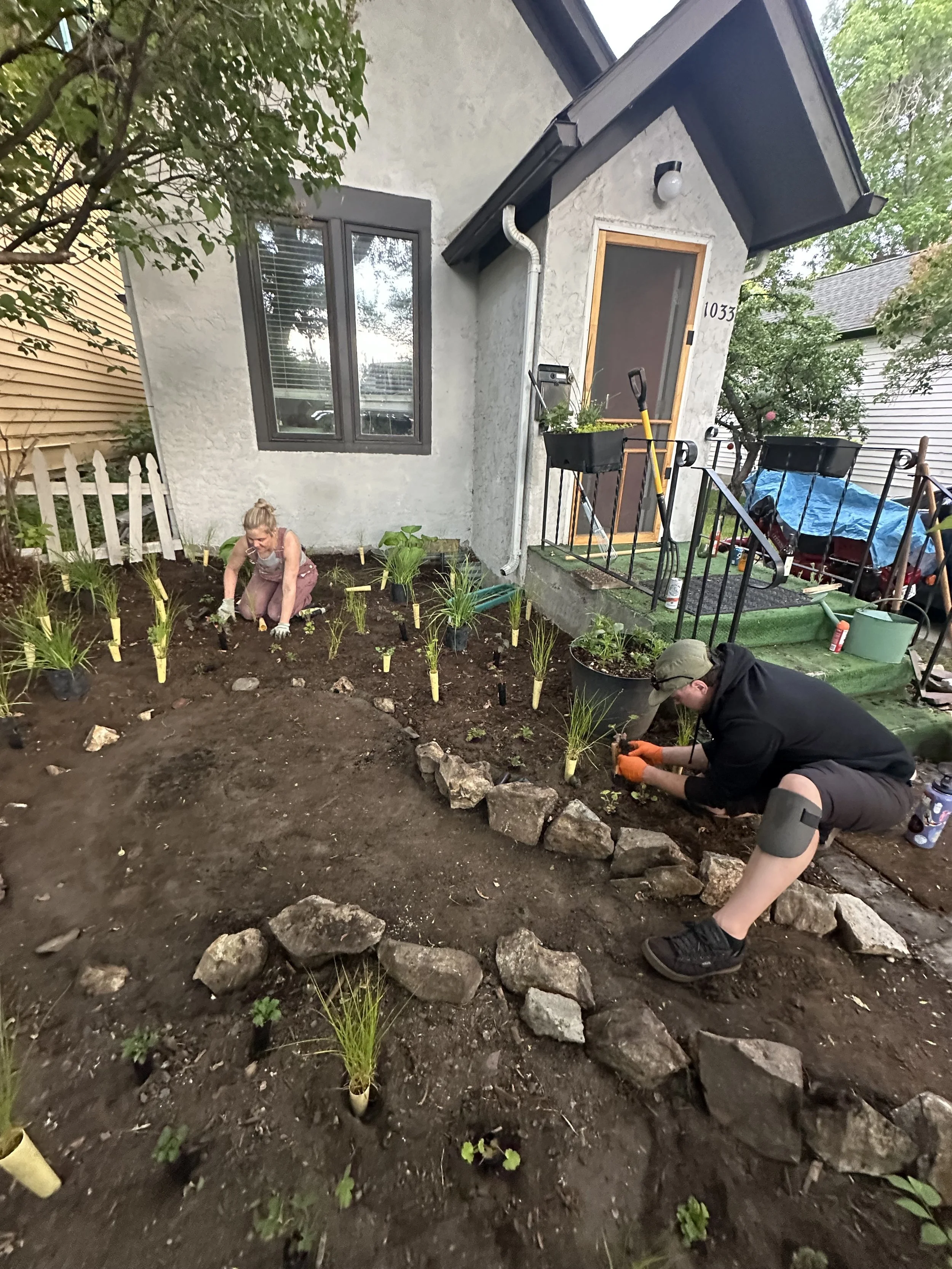 Two people planting flowers in a garden bed in front of a house with a white facade, black window frames, and a black screen door.