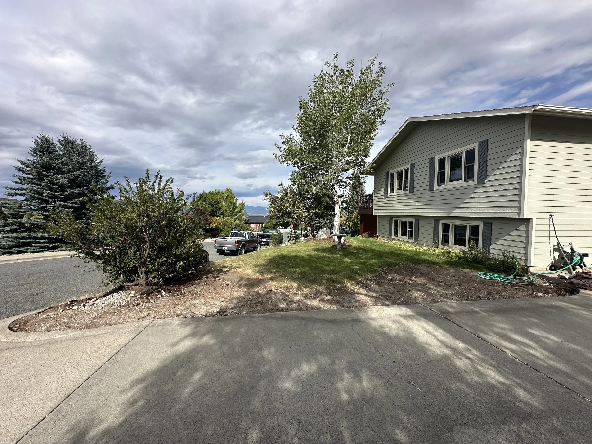 Residential neighborhood with a gray house, trees, parked cars, and a cloudy sky.