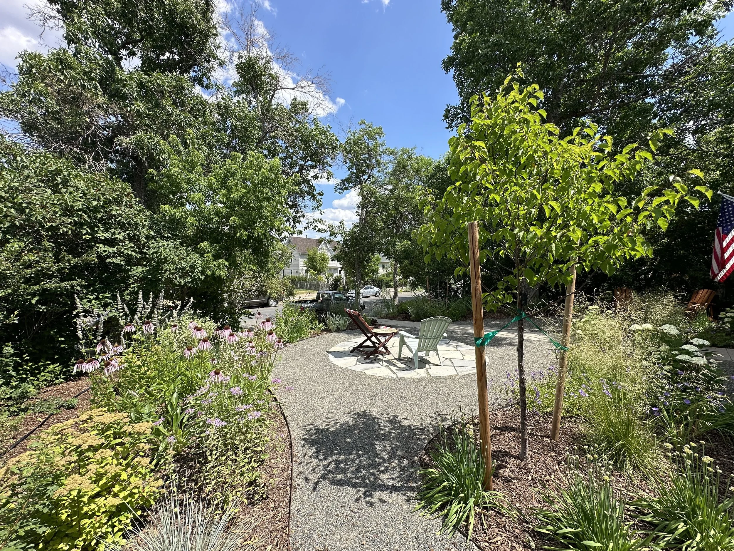 A garden with trees, flowering plants, and a outdoor seating area with two chairs on a paved surface, under a bright blue sky with a few clouds.