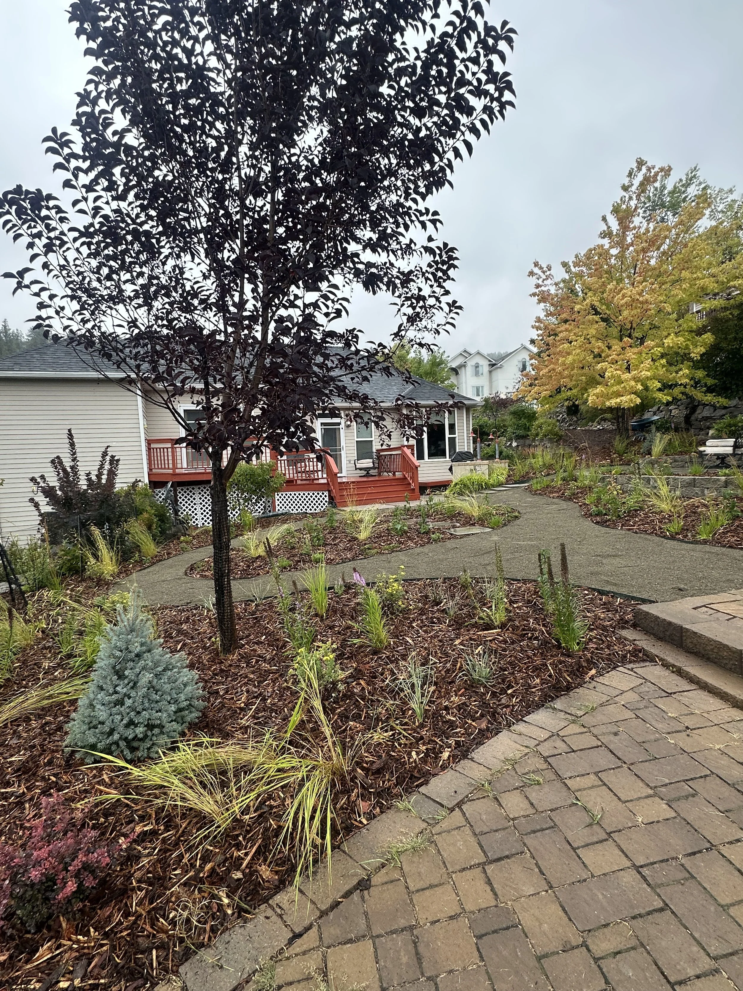 A landscaped backyard garden with a brick pathway, a wooden deck attached to a house, and trees with green and yellow leaves on a cloudy day.