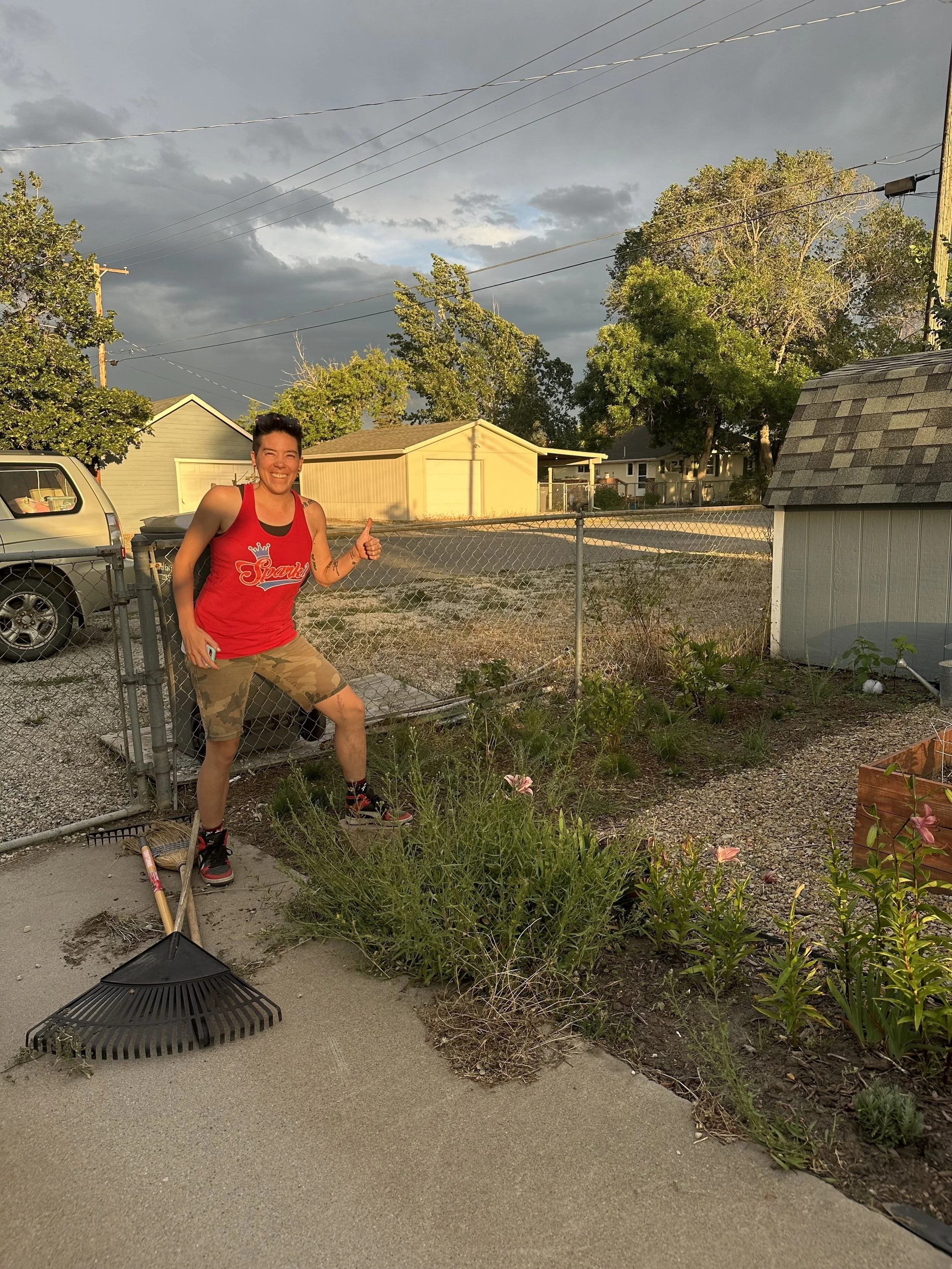 A woman smiling and giving a thumbs-up, standing outdoors near a garden, with a rake and shovel on the ground beside her, during sunset.