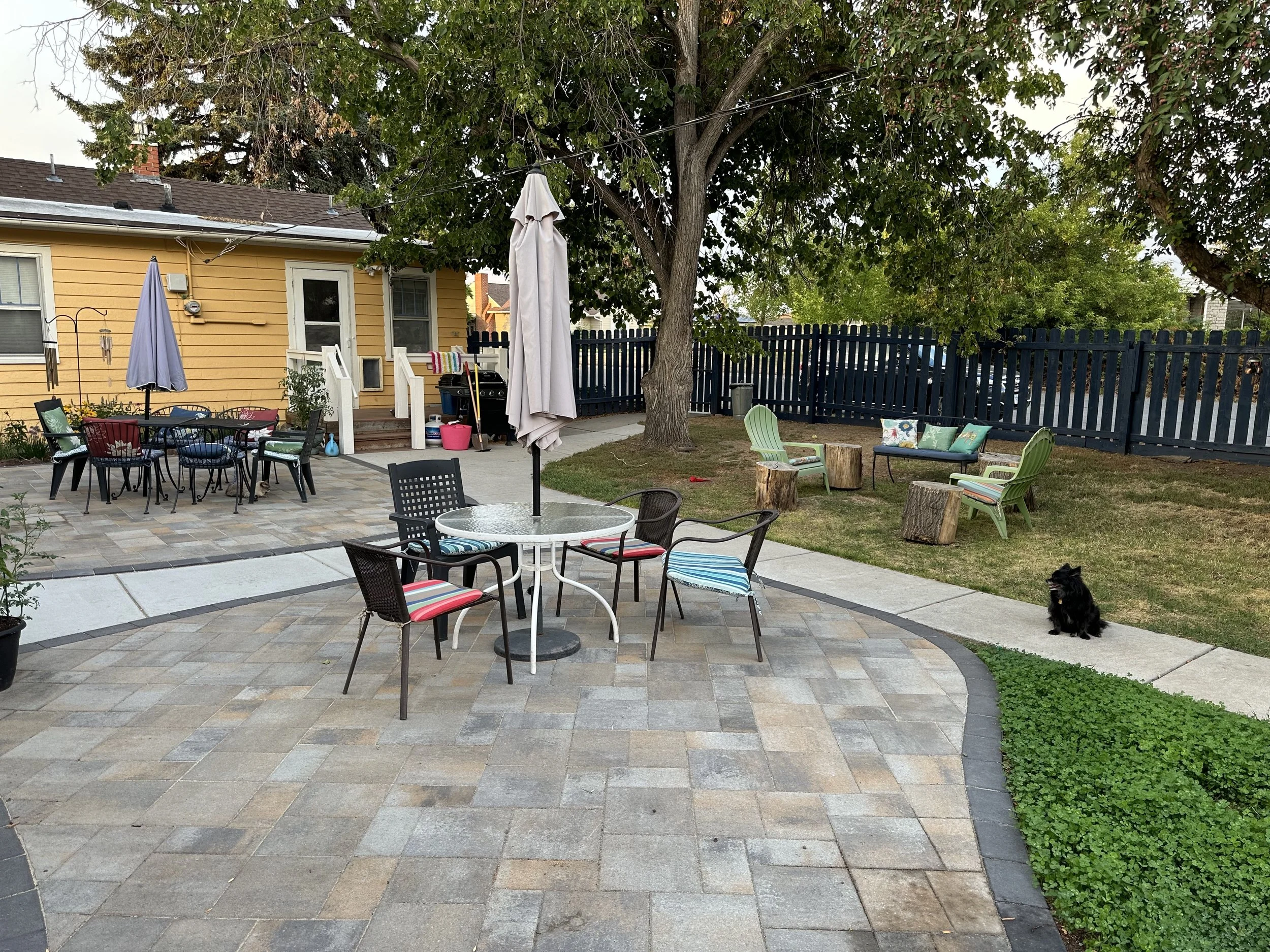 Backyard patio with outdoor furniture, including a round table with umbrellas, a rectangular table with multiple chairs, and a seating area with wooden logs and chairs near a large tree. A dog is sitting on the grass, and the yard is enclosed with a 