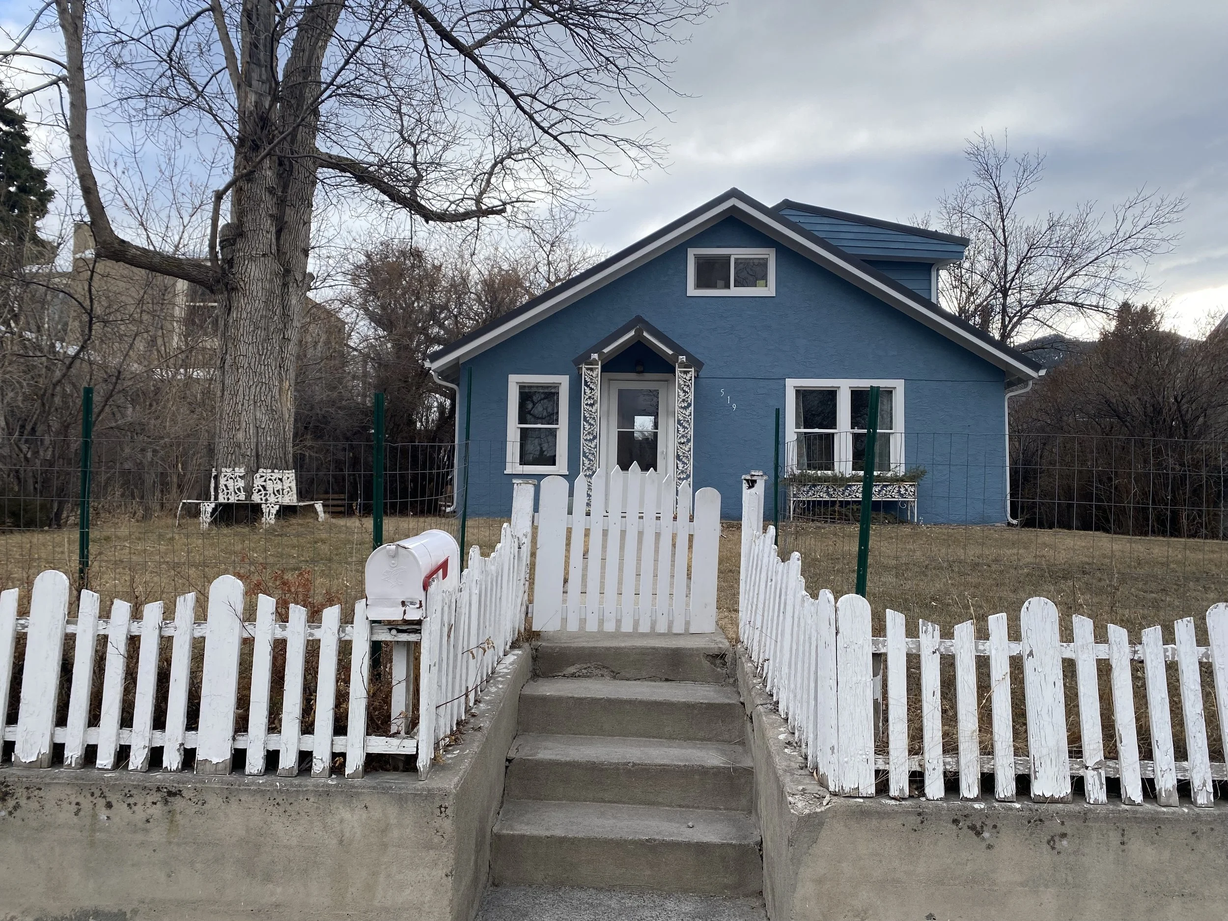 A blue house with white trim and a gabled roof, surrounded by a white picket fence and a concrete staircase leading to the front door, with a mailbox on the left side of the steps.