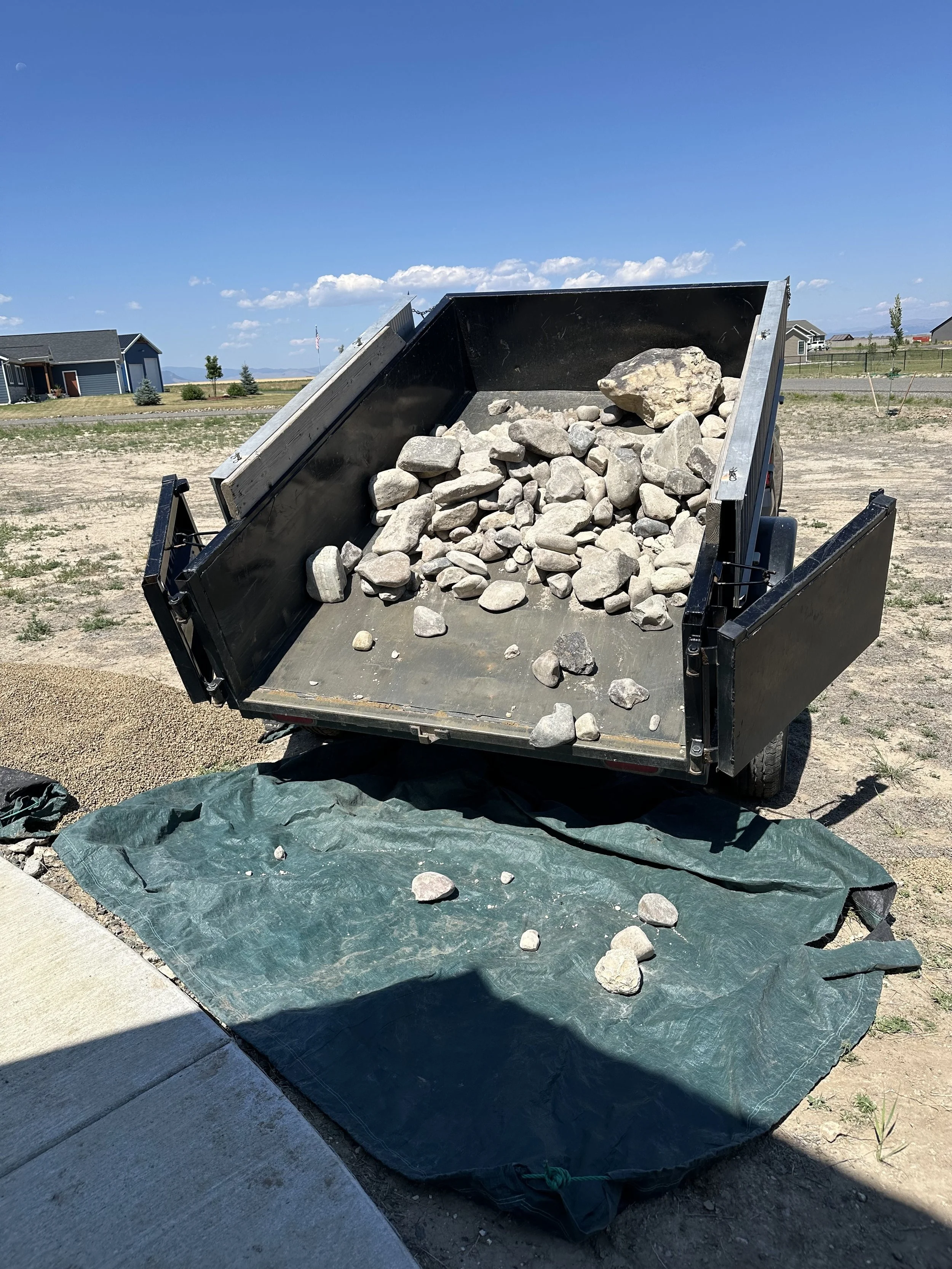 A dump trailer filled with rocks and gravel parked on a dirt lot, with a green tarp laid out underneath. Residential houses can be seen in the background under a blue sky.