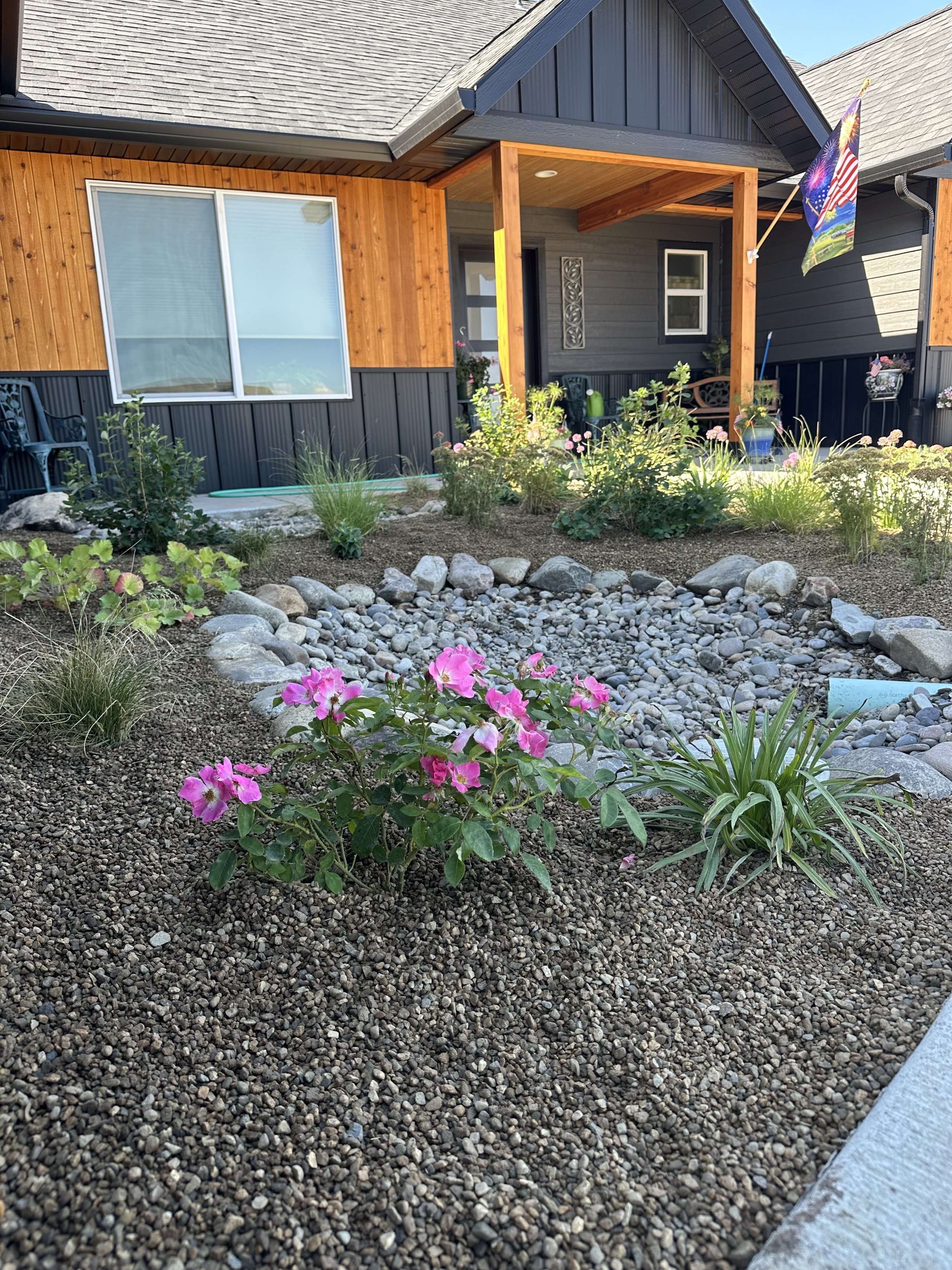 Front yard garden with pink flowers, small rocks, and a porch with a flag on a house.