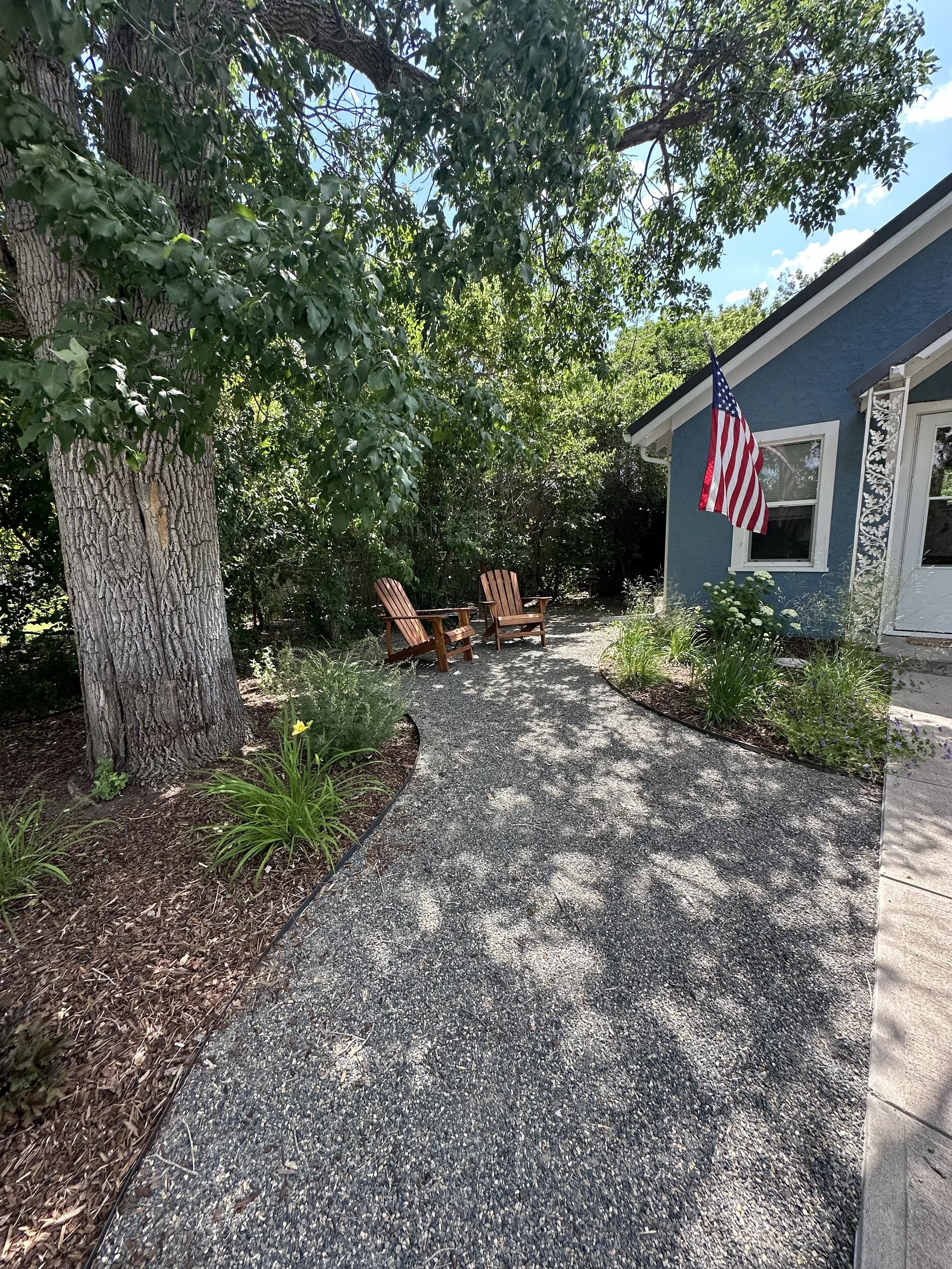 A gravel yard with two wooden chairs next to a large tree, beside a blue house with white trim and an American flag hanging on the wall. There are green plants and flowers in the garden beds along the house and a partly cloudy sky overhead.