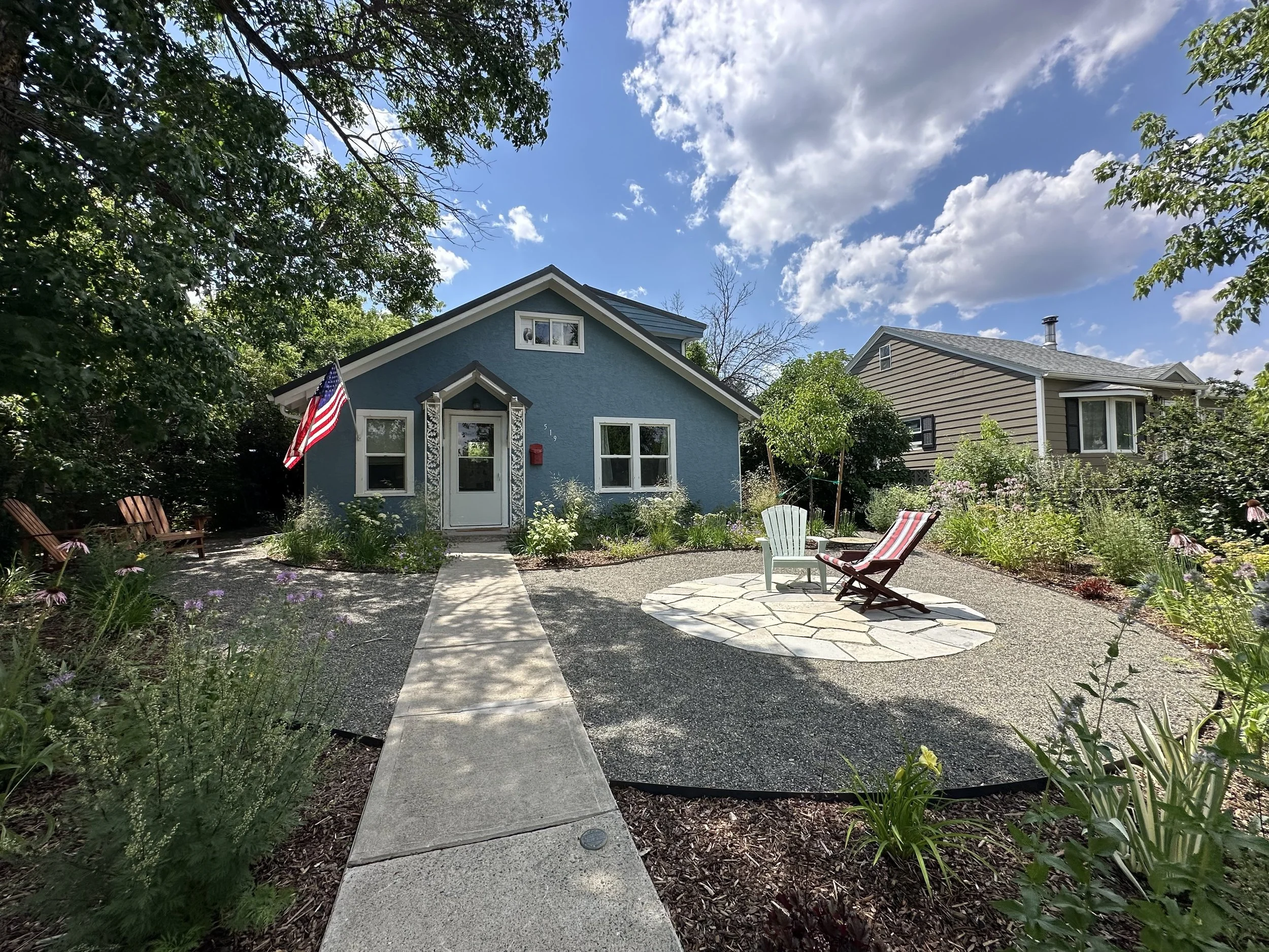 Front yard of a blue house with a concrete walkway leading to the front door, surrounded by a garden with colorful flowers and trees. There are three lawn chairs arranged around a small stone patio, and an American flag displayed near the house. The 