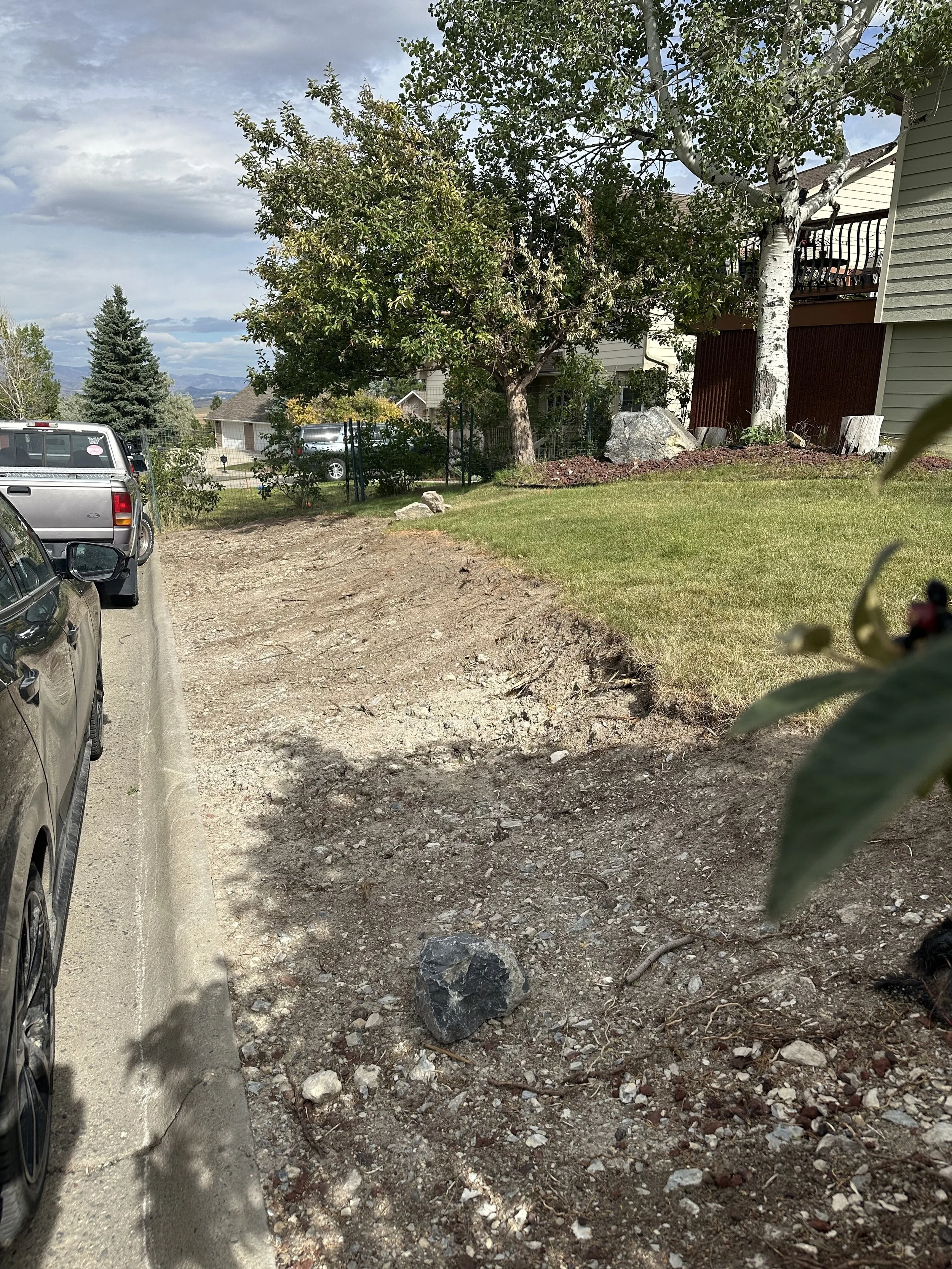 A partially cleared dirt area next to parked cars in a residential neighborhood, with grass, trees, and houses in the background.