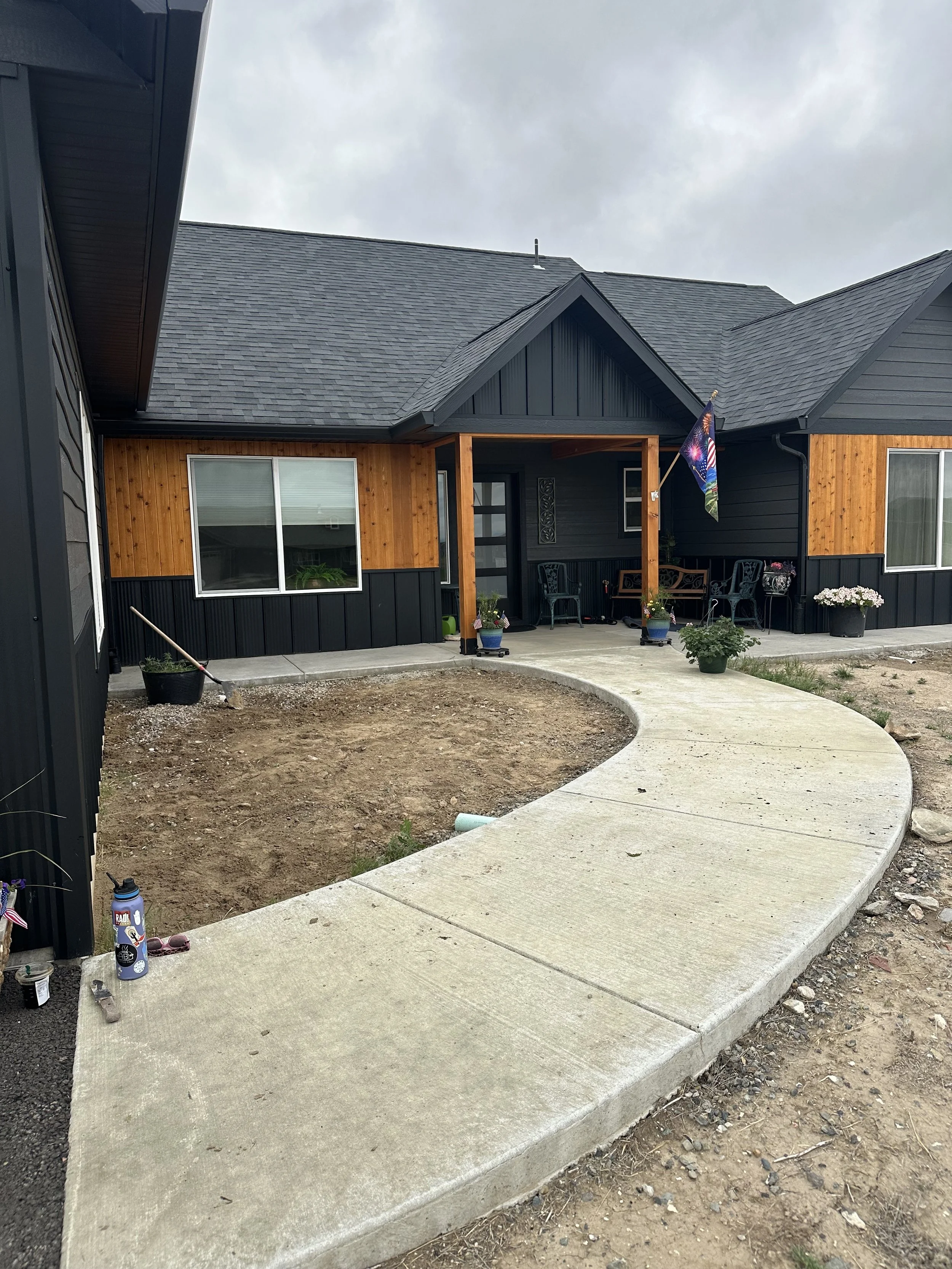 Front yard of a house under construction with a curved concrete walkway, potted plants, and patio furniture, with cloudy sky overhead.