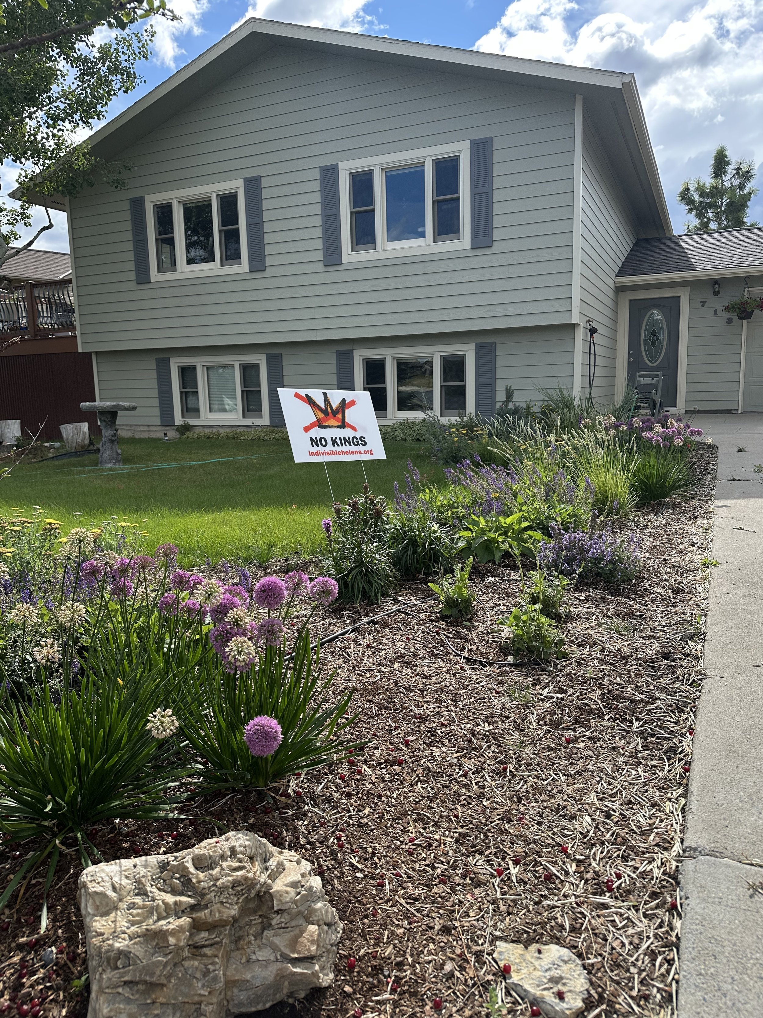 A front yard with a flower bed, rocks, and a lawn. A sign reads "No Kings" with a crossed-out crown symbol. A house with light gray siding and purple shutters in the background.