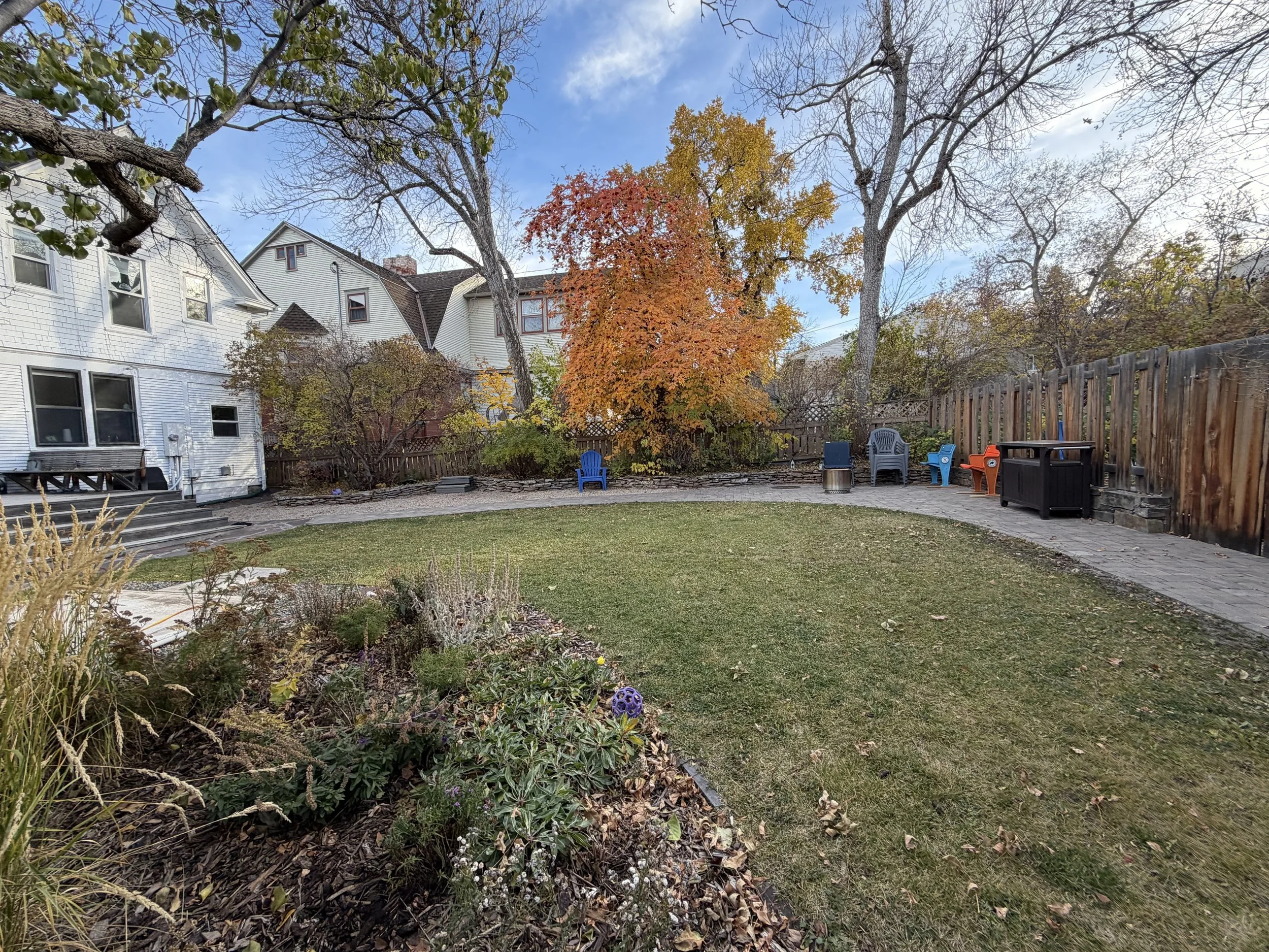A backyard with a grassy lawn and a paved patio area, surrounded by trees with autumn leaves, and colorful outdoor chairs along a wooden fence.