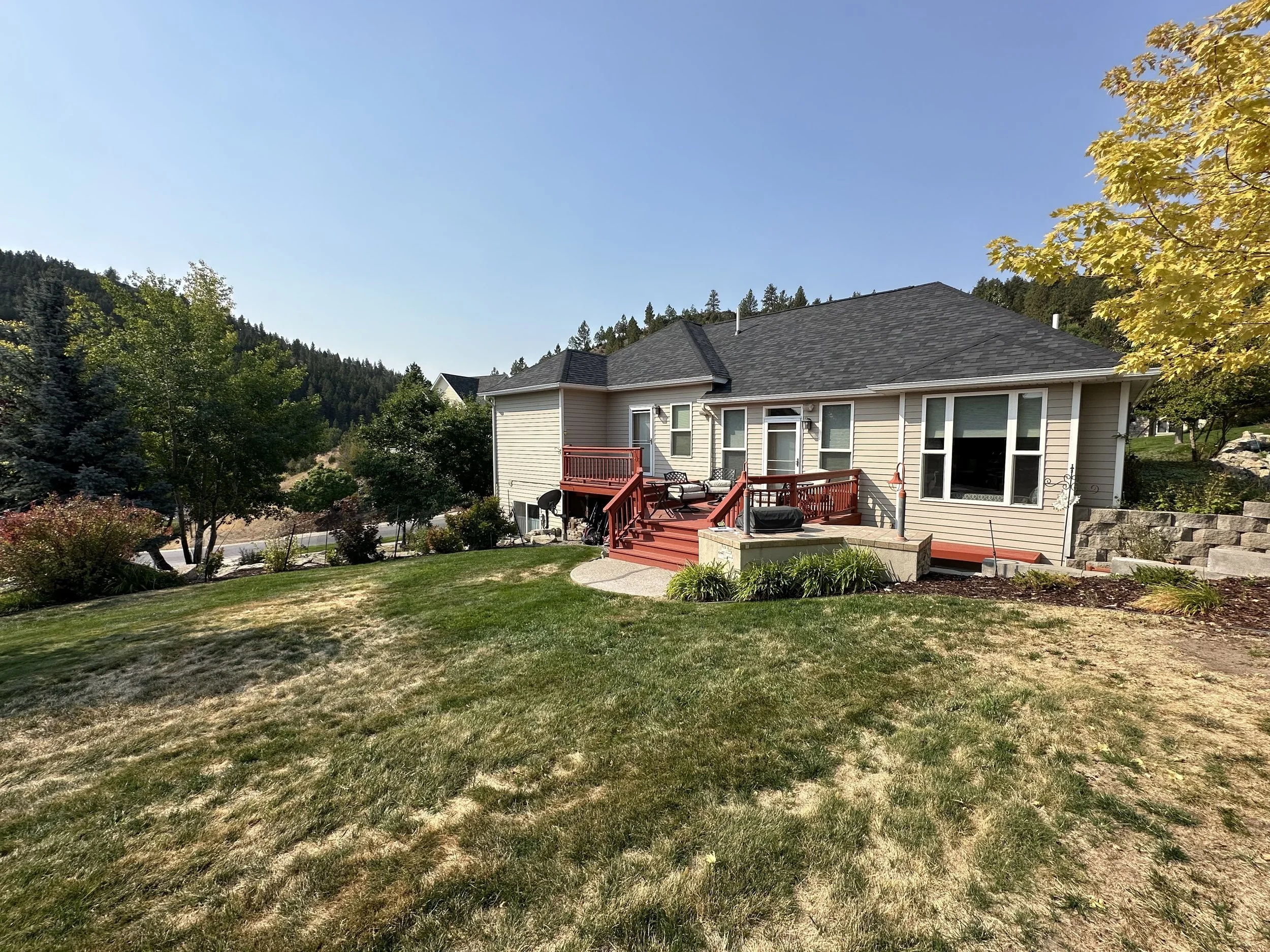 A backyard with a beige house featuring a red deck, surrounded by greenery and trees, with rolling hills and mountains in the background under a clear blue sky.