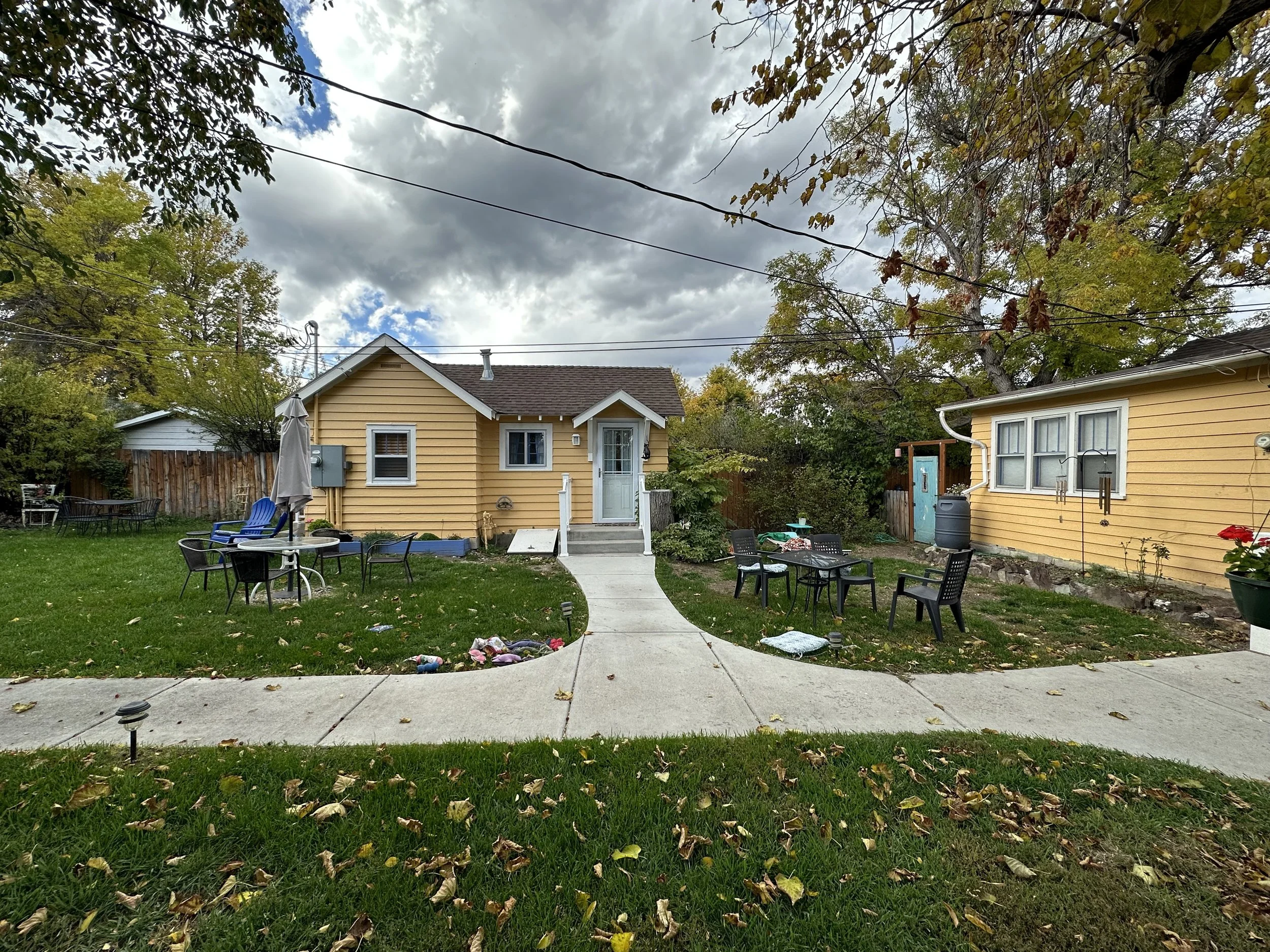 A backyard with a concrete walkway, outdoor furniture, and a yellow house with white trim on a cloudy day.
