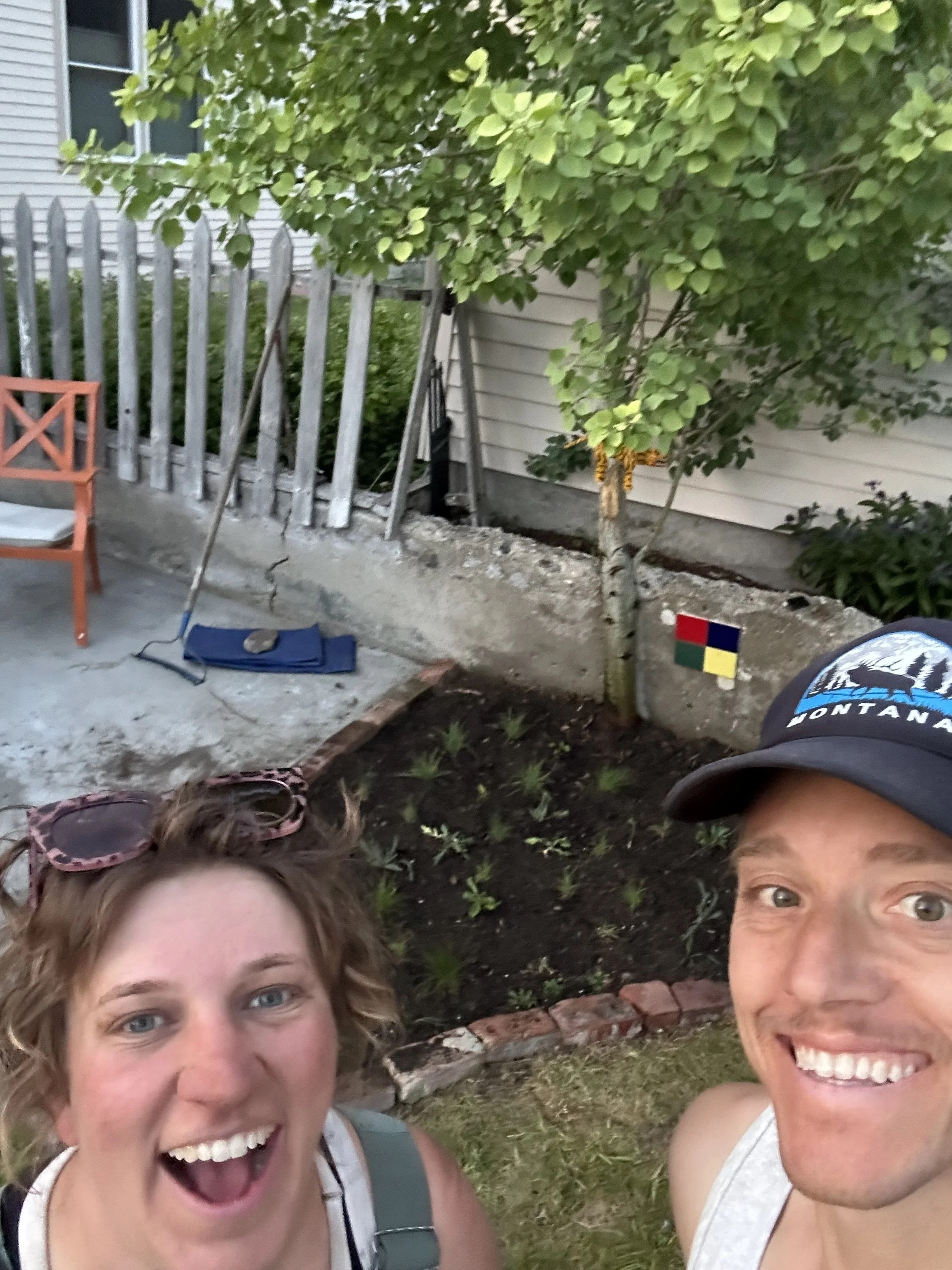 A smiling woman and man taking a selfie in front of a garden bed with a small tree and young plants, a painted rainbow icon on the wall, and a partially visible fence and house in the background.