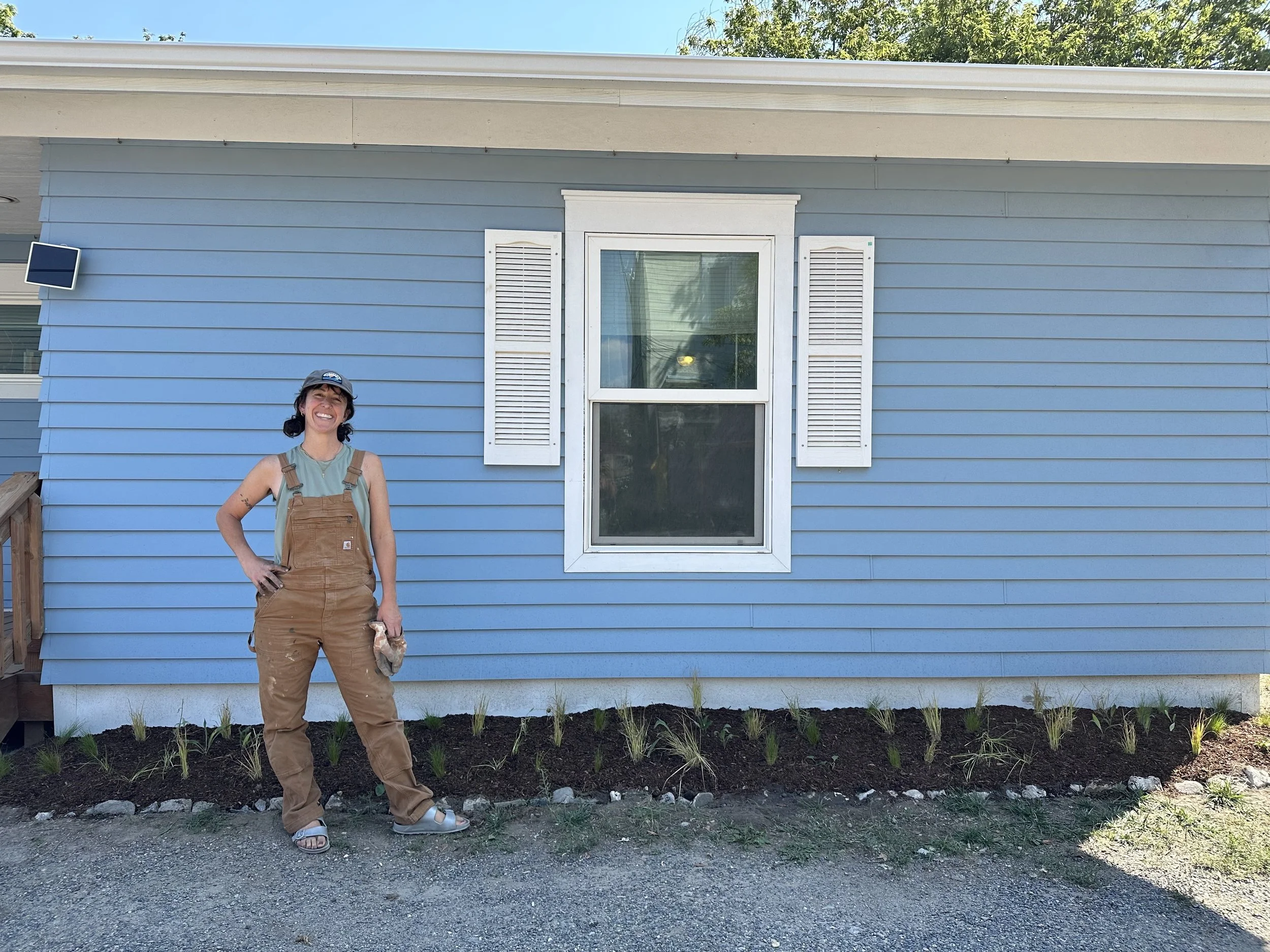 A person standing in front of a blue house with white window shutters, wearing brown work overalls, a grey t-shirt, a baseball cap, and sandals, smiling, with a small garden in front.