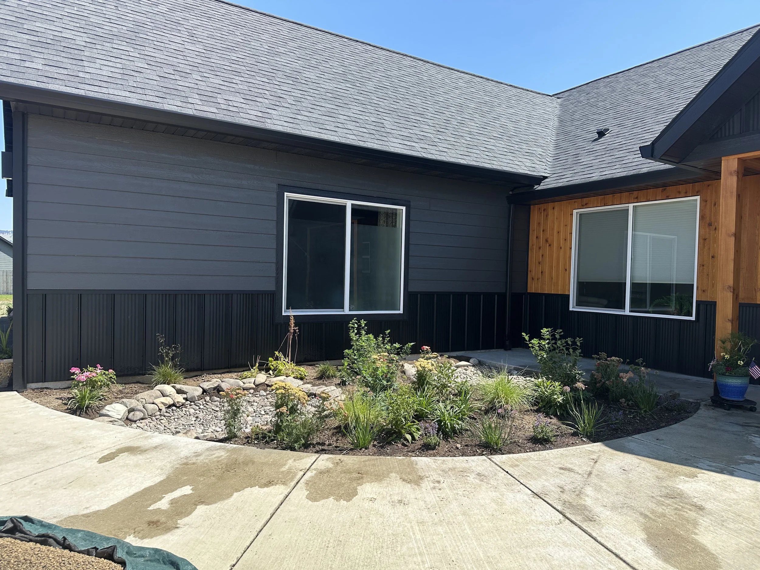 Exterior view of a house with gray siding, black trim, and a section of wood paneling, featuring two large glass windows and a garden bed with plants and flowers in front, under a clear blue sky.
