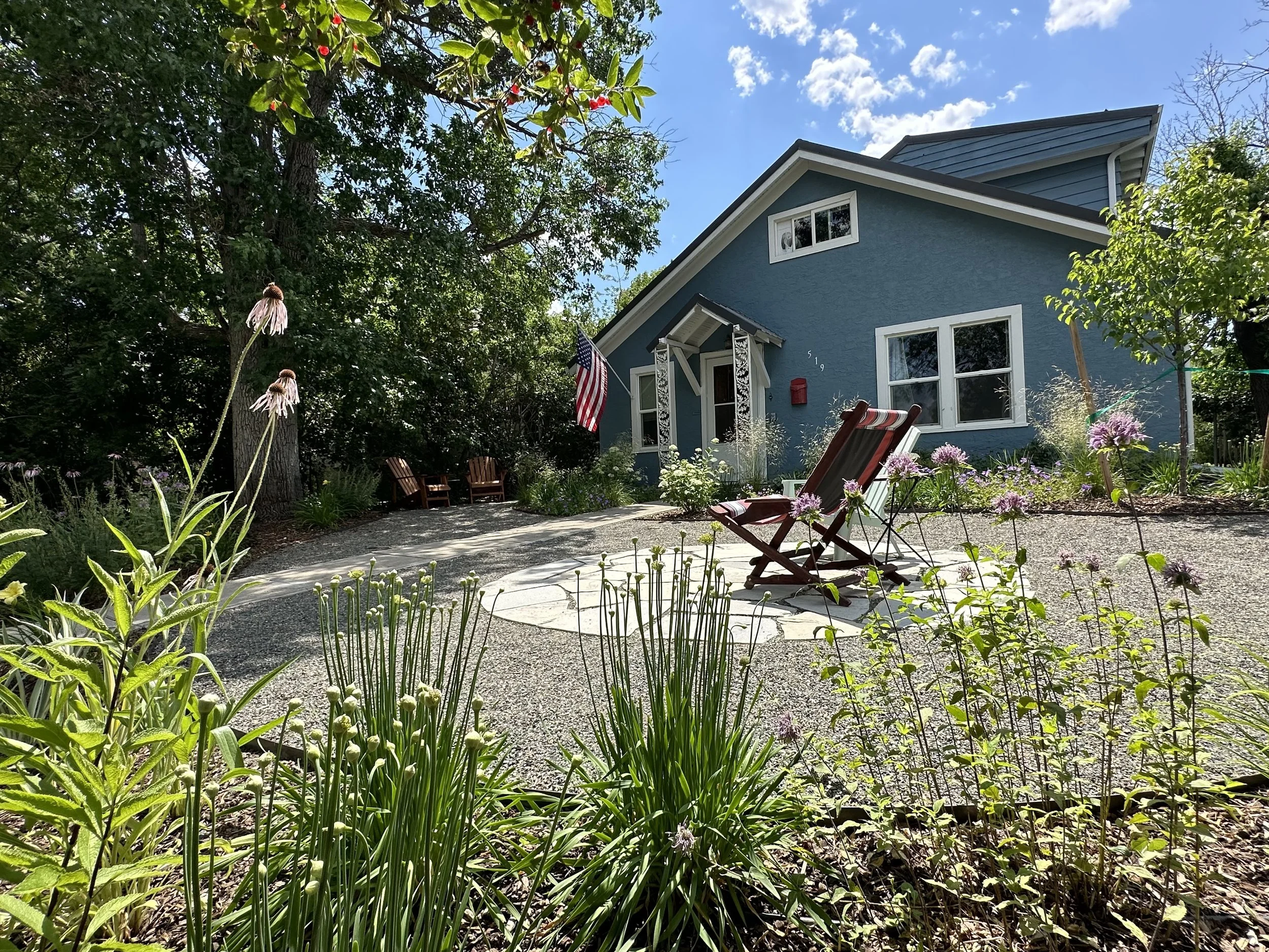 A blue house with white trim and a porch, surrounded by a lush garden with flowers, trees, and a gravel pathway. A wooden reclining chair and an American flag are visible in the yard, under a partly cloudy sky.