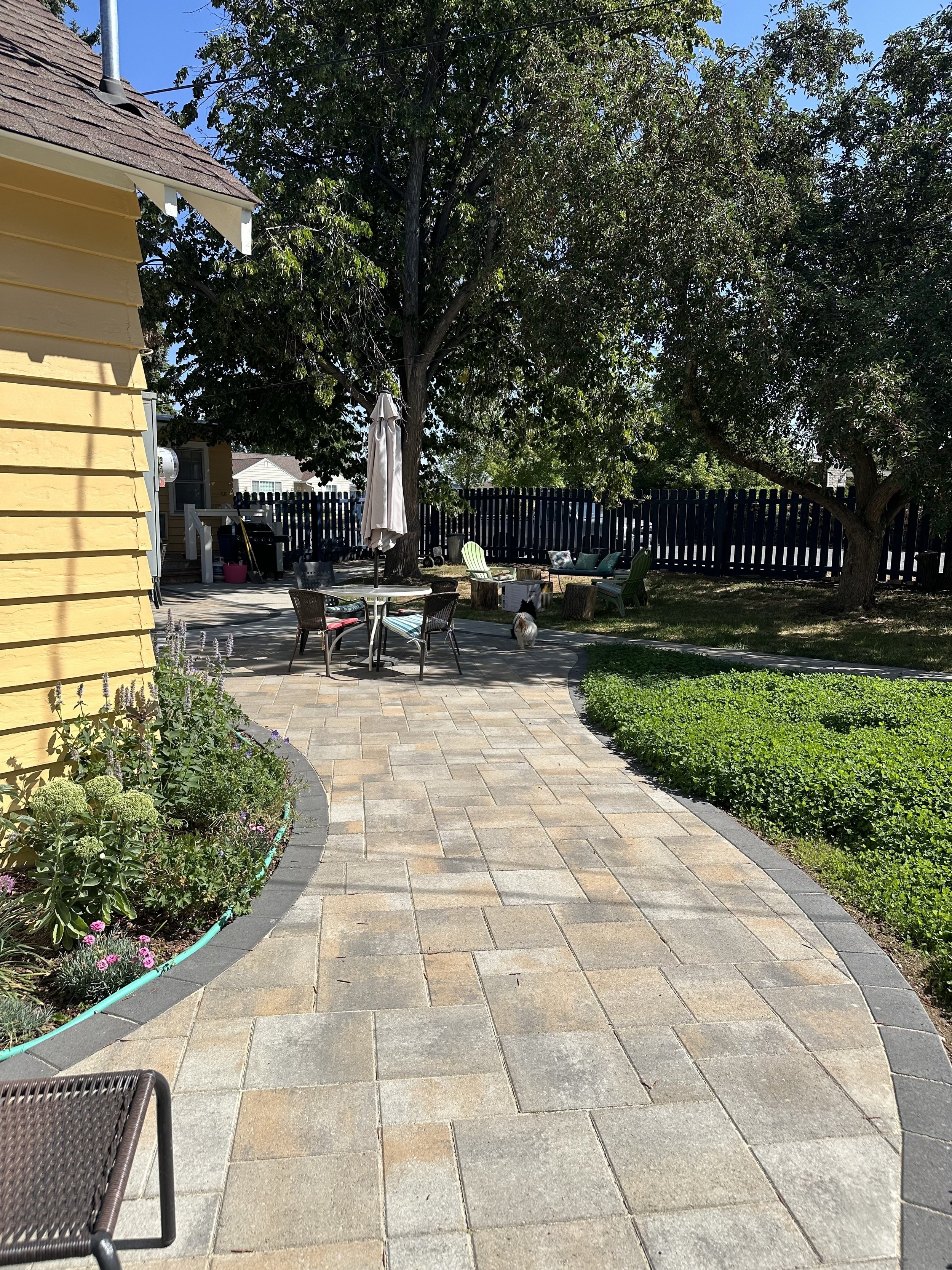 Backyard patio with a curved stone pathway, garden beds with flowers on the left, a table with an umbrella, several chairs, and a dog near the trees on the right, enclosed by a black fence.
