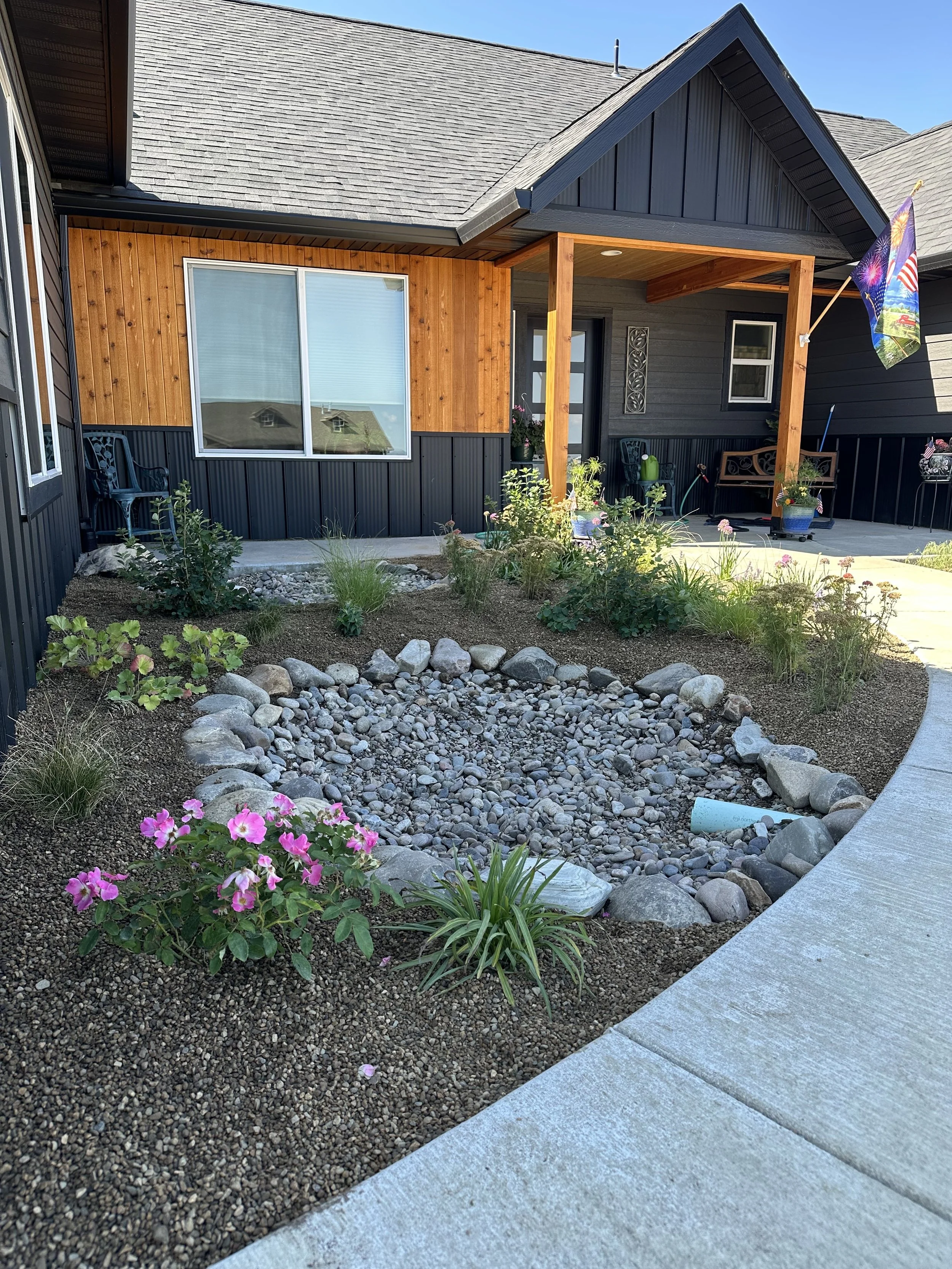 Front view of a house with a wooden and black siding exterior, a small porch with seating, a garden bed with pink flowers, green plants, rocks, and a flag on the porch.