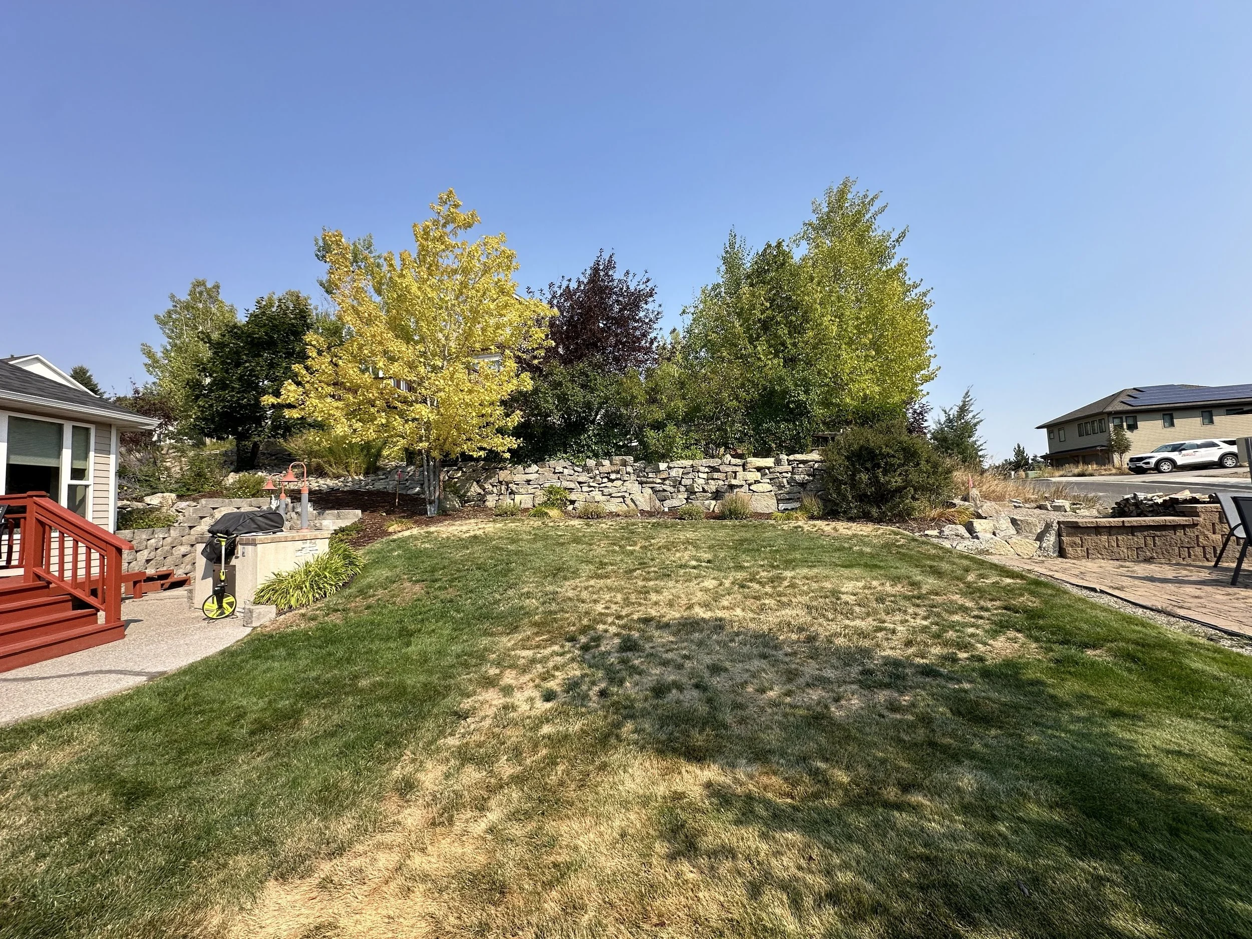 A backyard with a lawn, trees, stone retaining wall, and patio area with outdoor furniture under a clear blue sky.