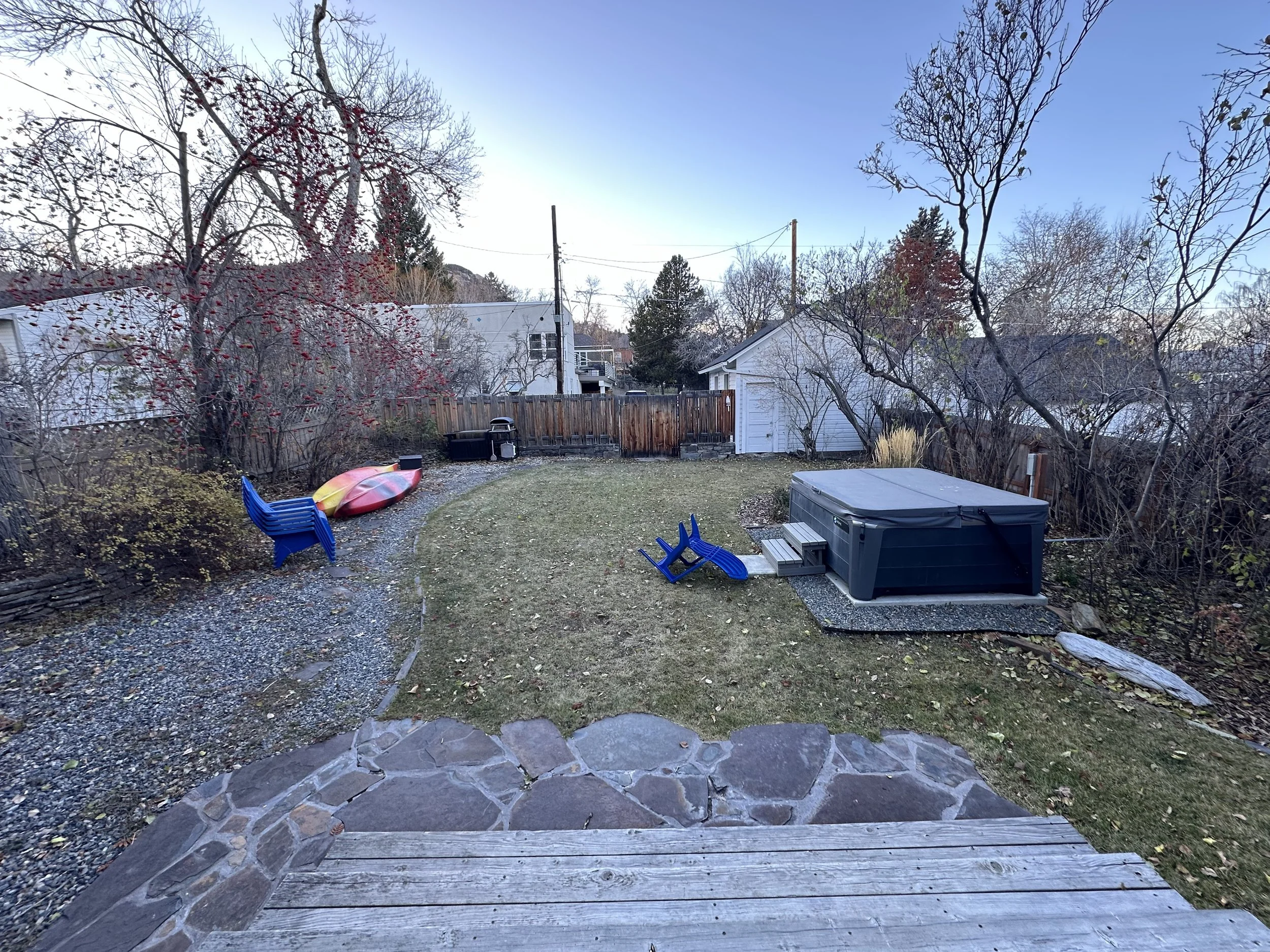 Backyard with a stone patio, grass lawn, leafless trees, and backyard toys including a blue chair and sled, a hot tub, and kayaks, during late fall or early winter.