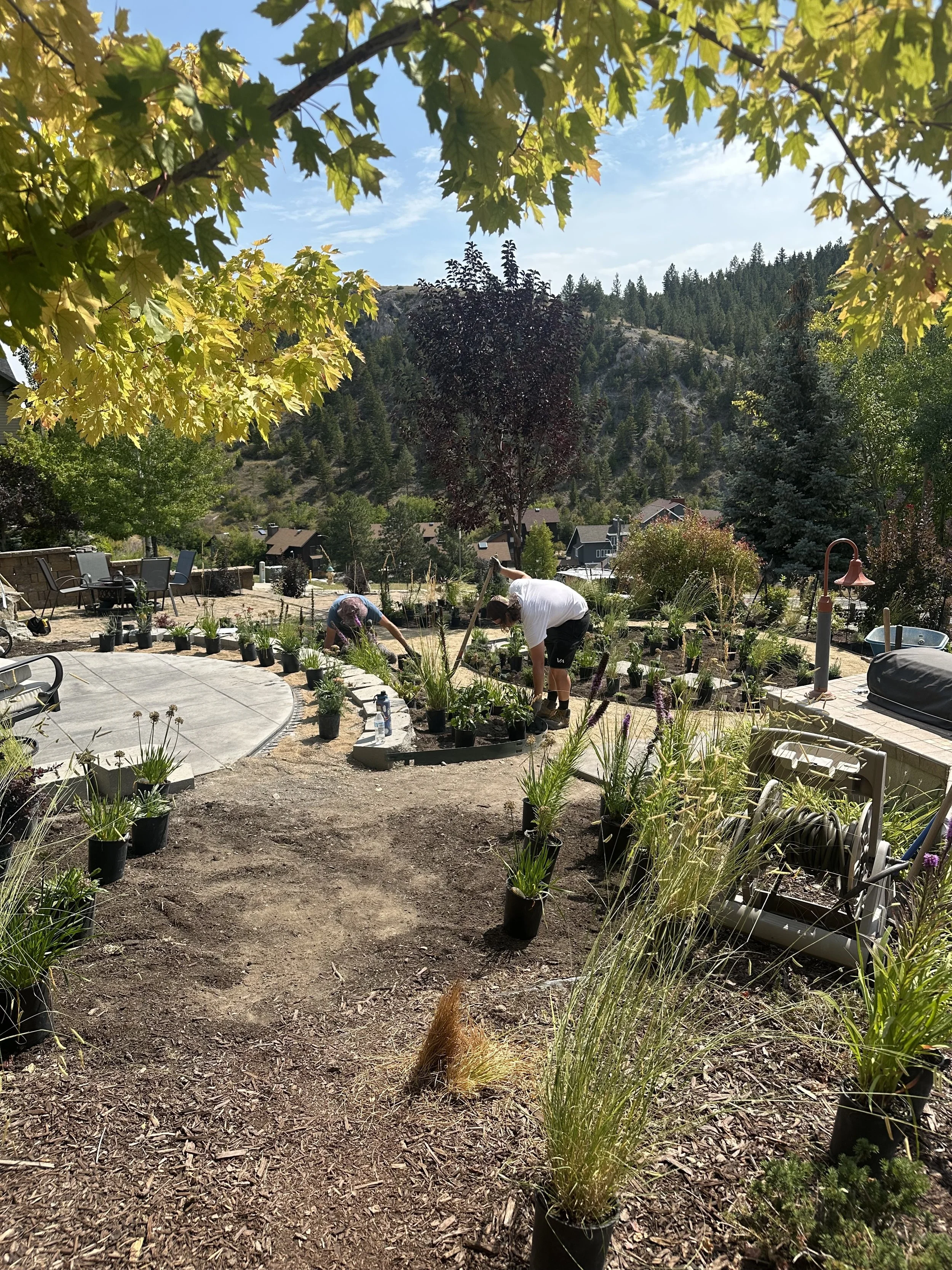 Two people working in a garden, planting and tending to plants in black pots. The garden is on a hillside with scenic mountain views in the background. Bright sunny day with clear skies, trees, and outdoor furniture visible.