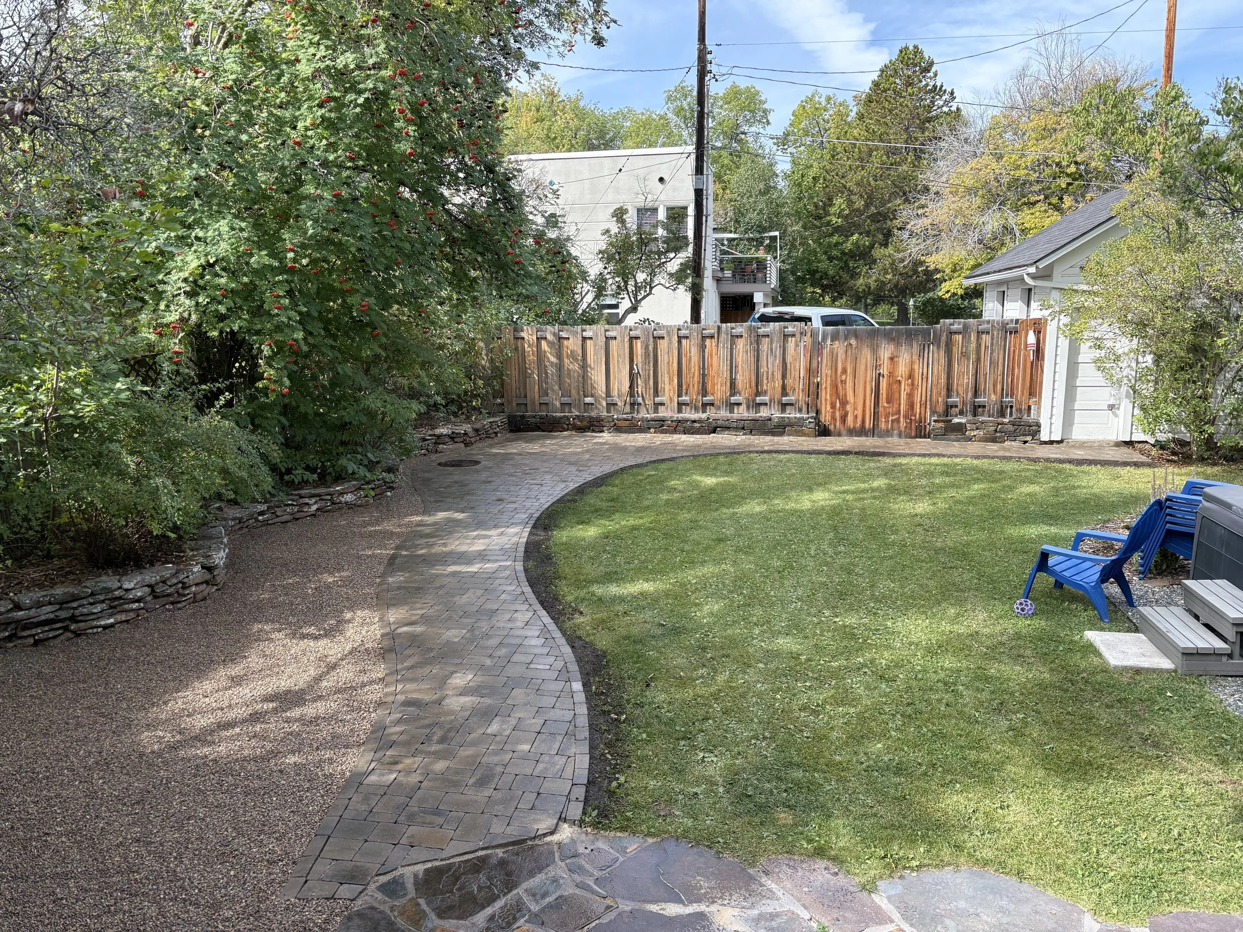 A backyard with a curving stone and brick pathway, a grassy lawn, a wooden fence, a small shed, and stacked blue chairs on the right.