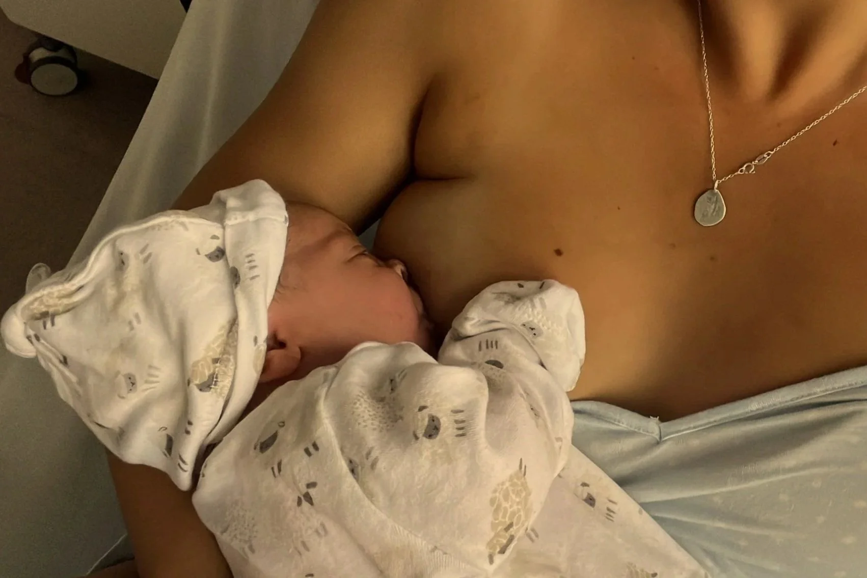 a newborn baby feeding at the breast, dressed in a matching grow and hat, mother wearing a silver necklace