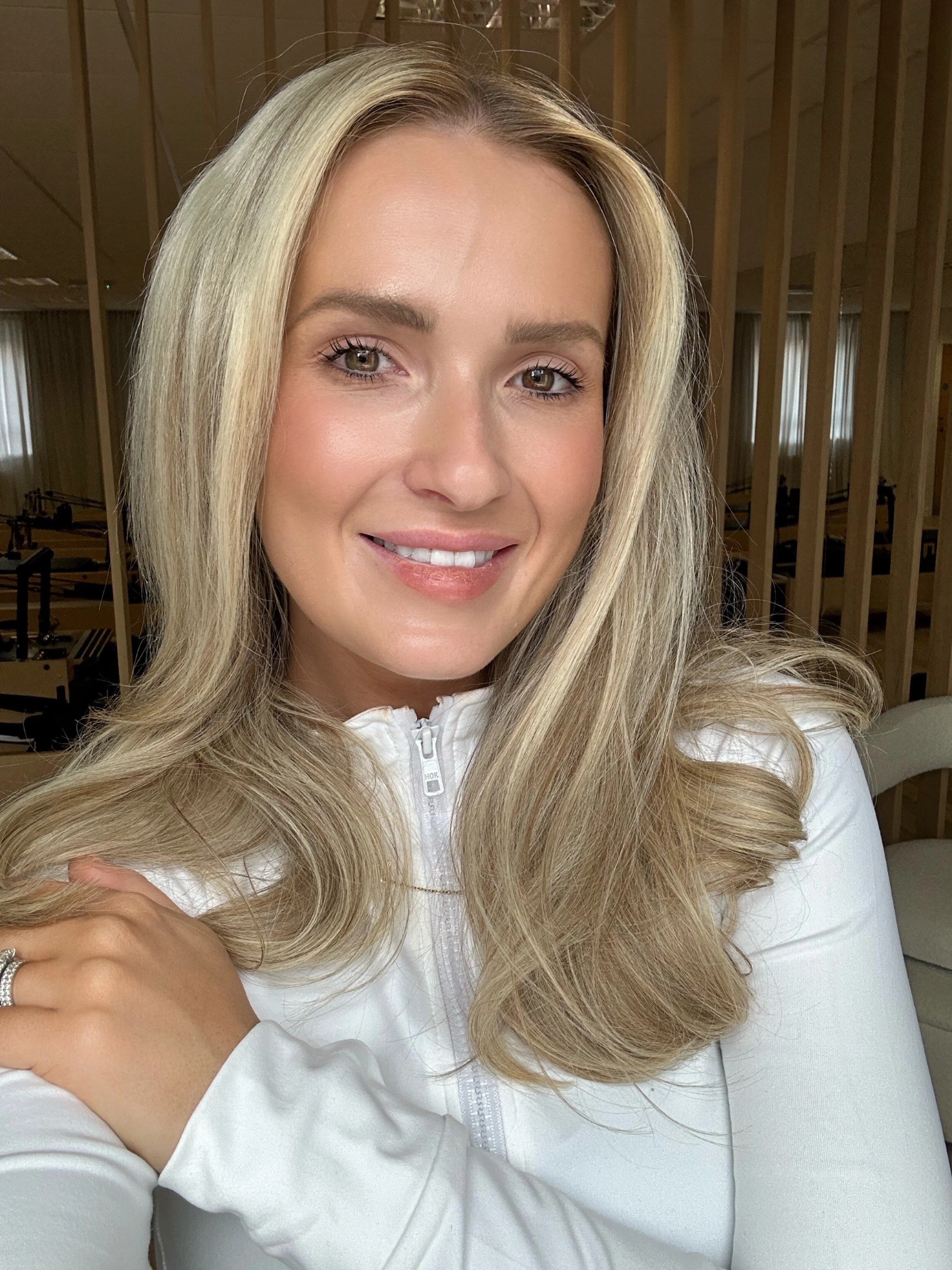 Woman with long blonde hair wearing a white zip-up top, smiling at the camera in an indoor setting.