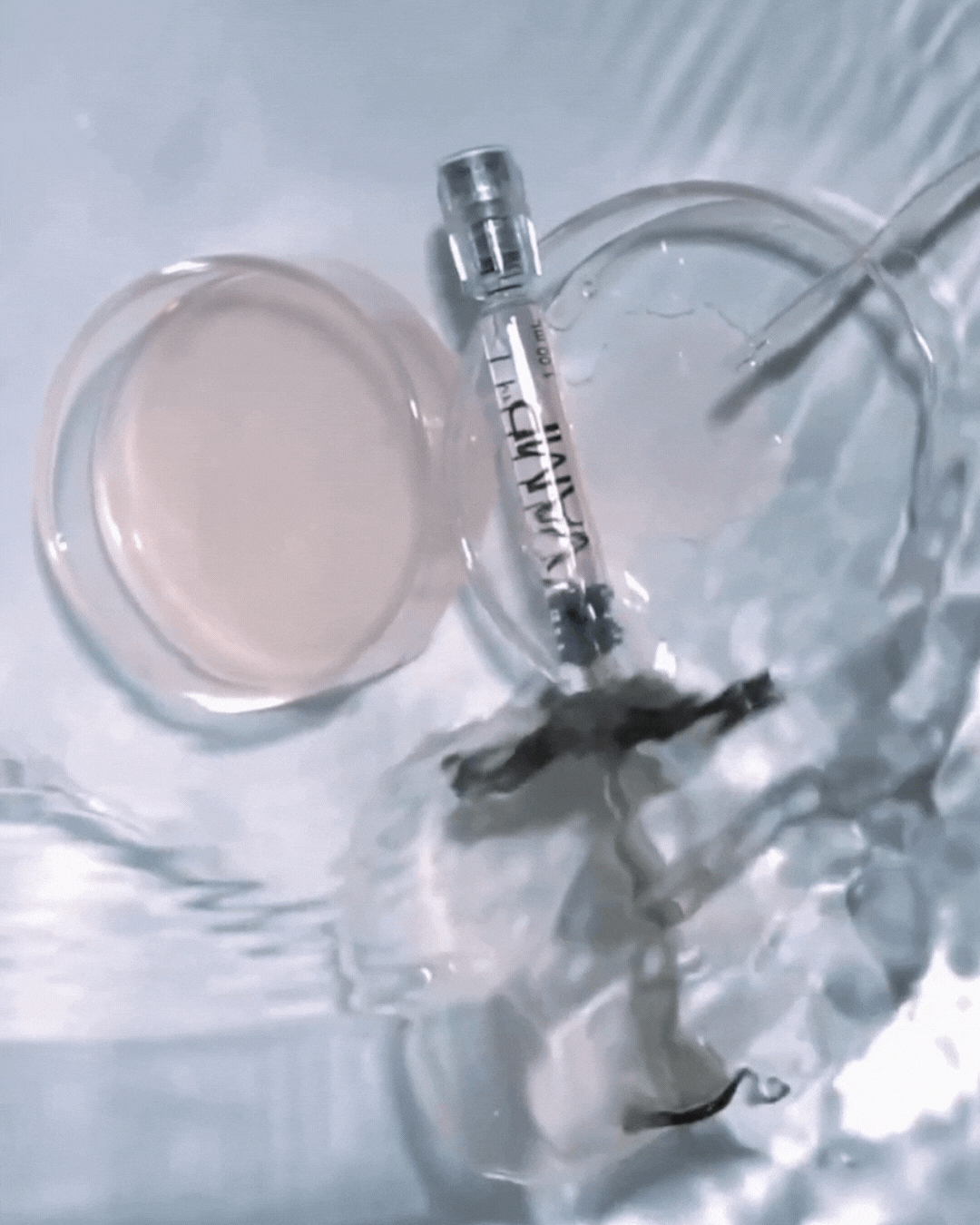 An injection syringe partially submerged in water, next to a water-filled container and a glass of water with ice, all on a light-colored surface.