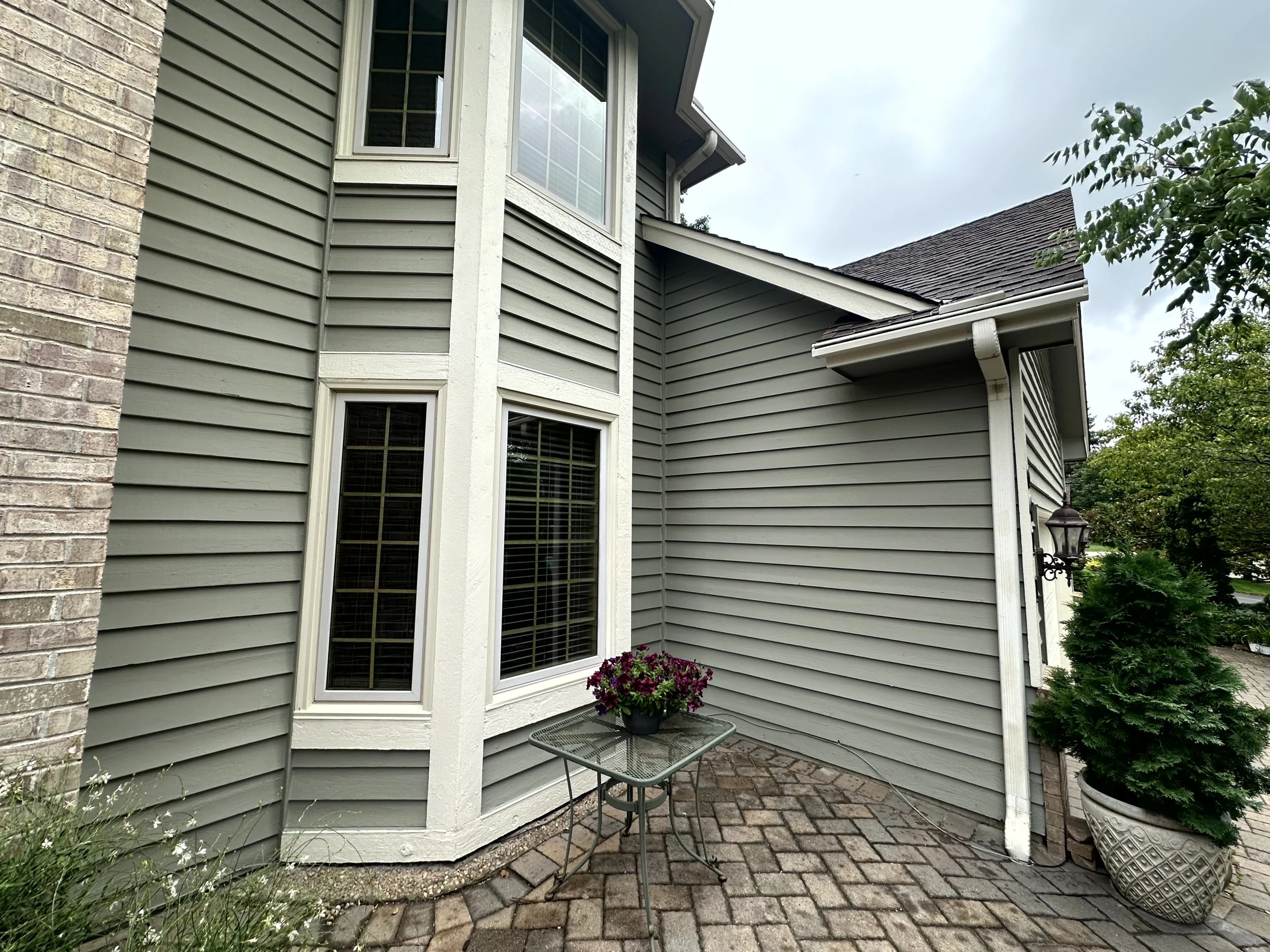 Outside corner of a house with green siding, white trim, and brick accents, featuring two windows, a small table with a potted flower, and a large potted plant nearby.