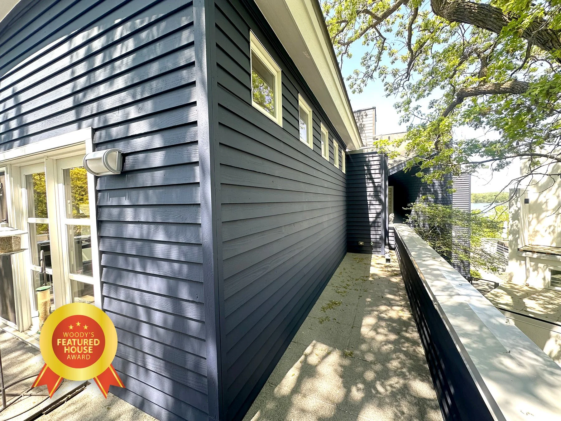 Modern building exterior with dark blue horizontal siding, several small high windows, a balcony with a black railing, and trees casting shadows on the sidewalk.