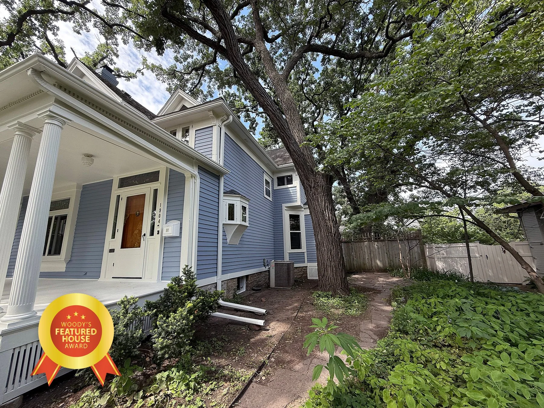 Blue house with white trim and porch, surrounded by trees and greenery, with a wooden fence in the backyard, featuring a 'Woody's Featured House Award' badge at the bottom left.