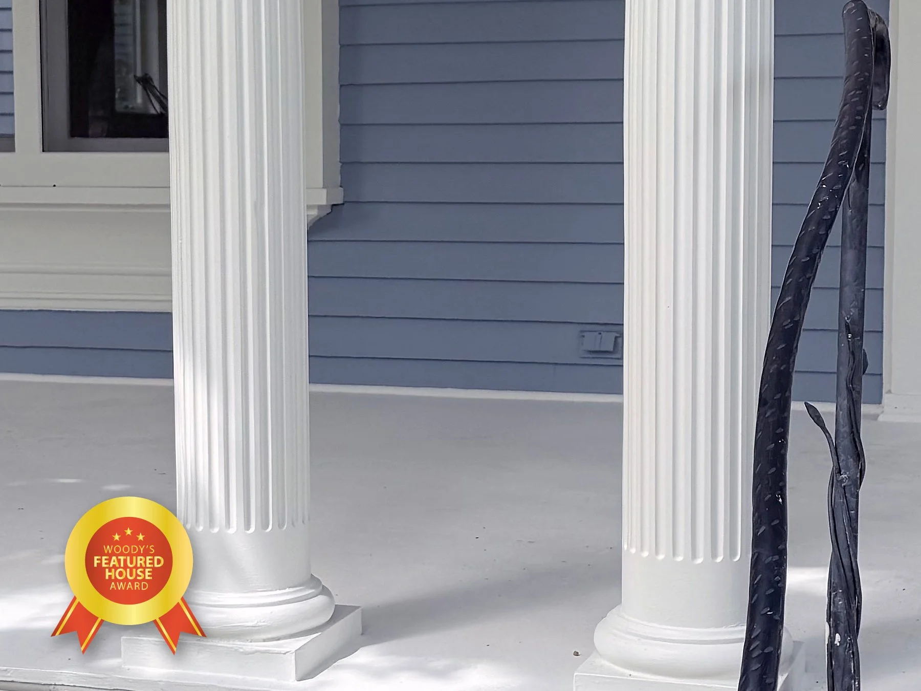 Close-up of two white decorative columns on a porch with blue siding in the background. There is a black cable or hose hanging on the right side and a small award badge on the bottom left corner that reads 'Woody's Featured House Award'.