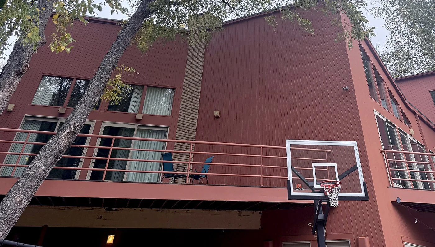 A multi-story building with red exterior siding, featuring several windows and a balcony with chairs. A basketball hoop is attached to the building's corner beneath the balcony. Trees with green leaves are in the foreground.