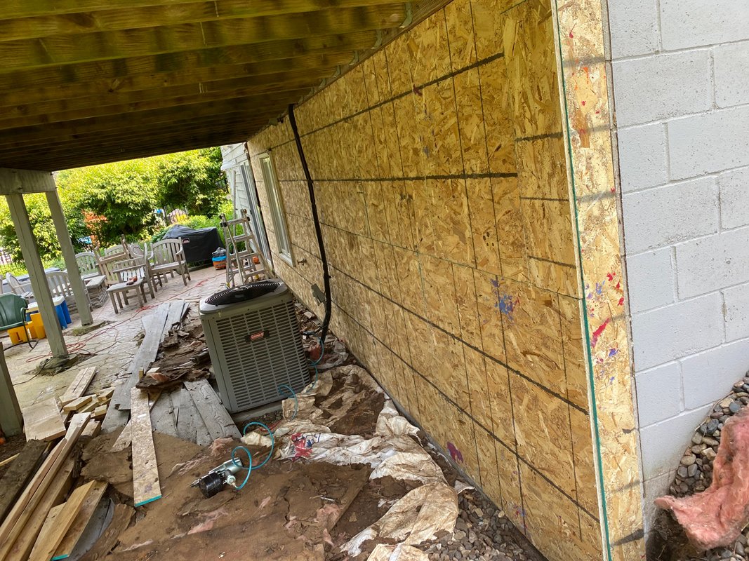 Side of a house under construction with exposed OSB sheathing, facing a porch with chairs and trees in the background, construction materials and equipment on the ground.