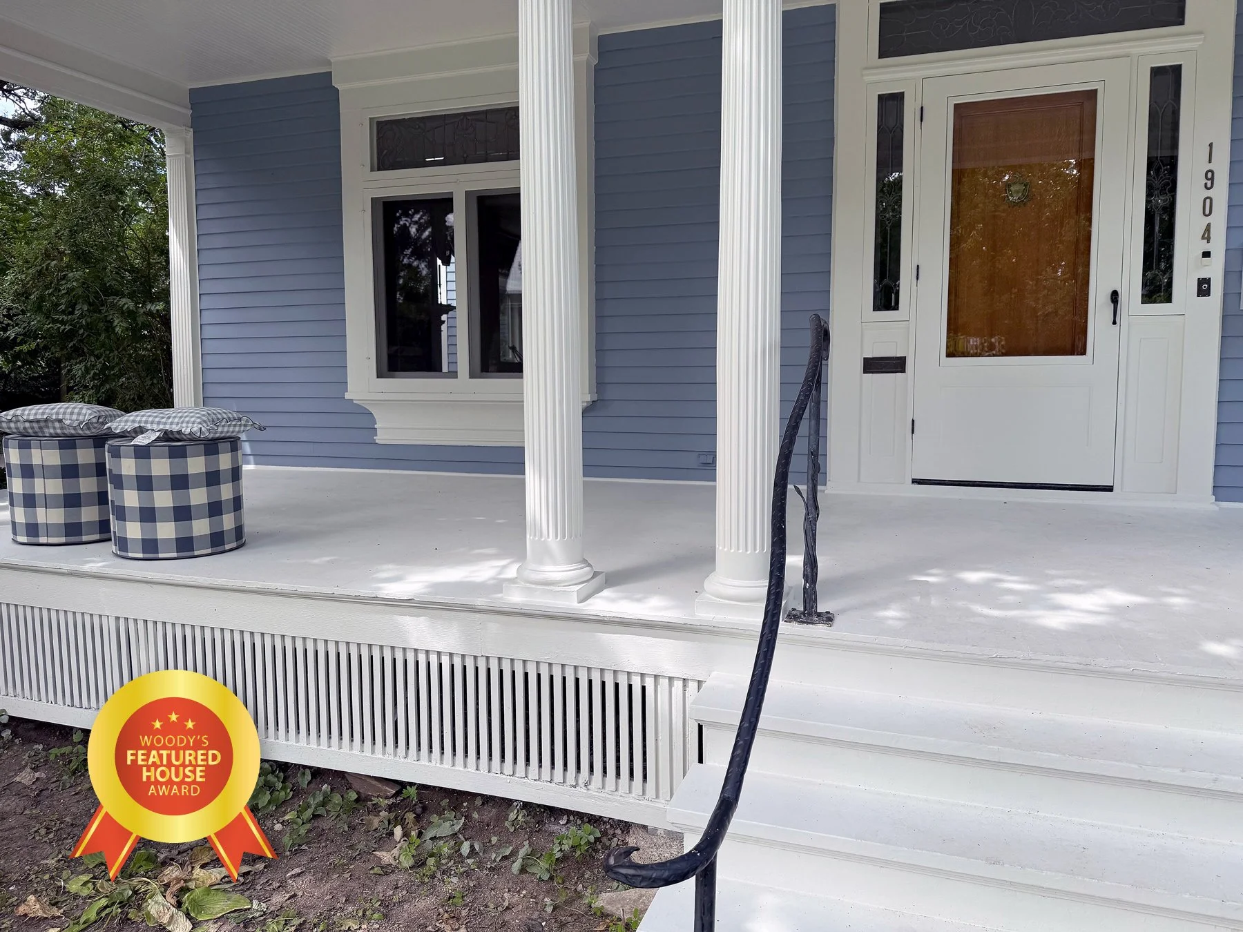 Front porch of a house with blue siding, white trim, white railing, and white columns. There are two plaid ottomans with pillows on top. There is a black handrail on the steps and a 'Woody's Featured House Award' badge at the bottom left corner.