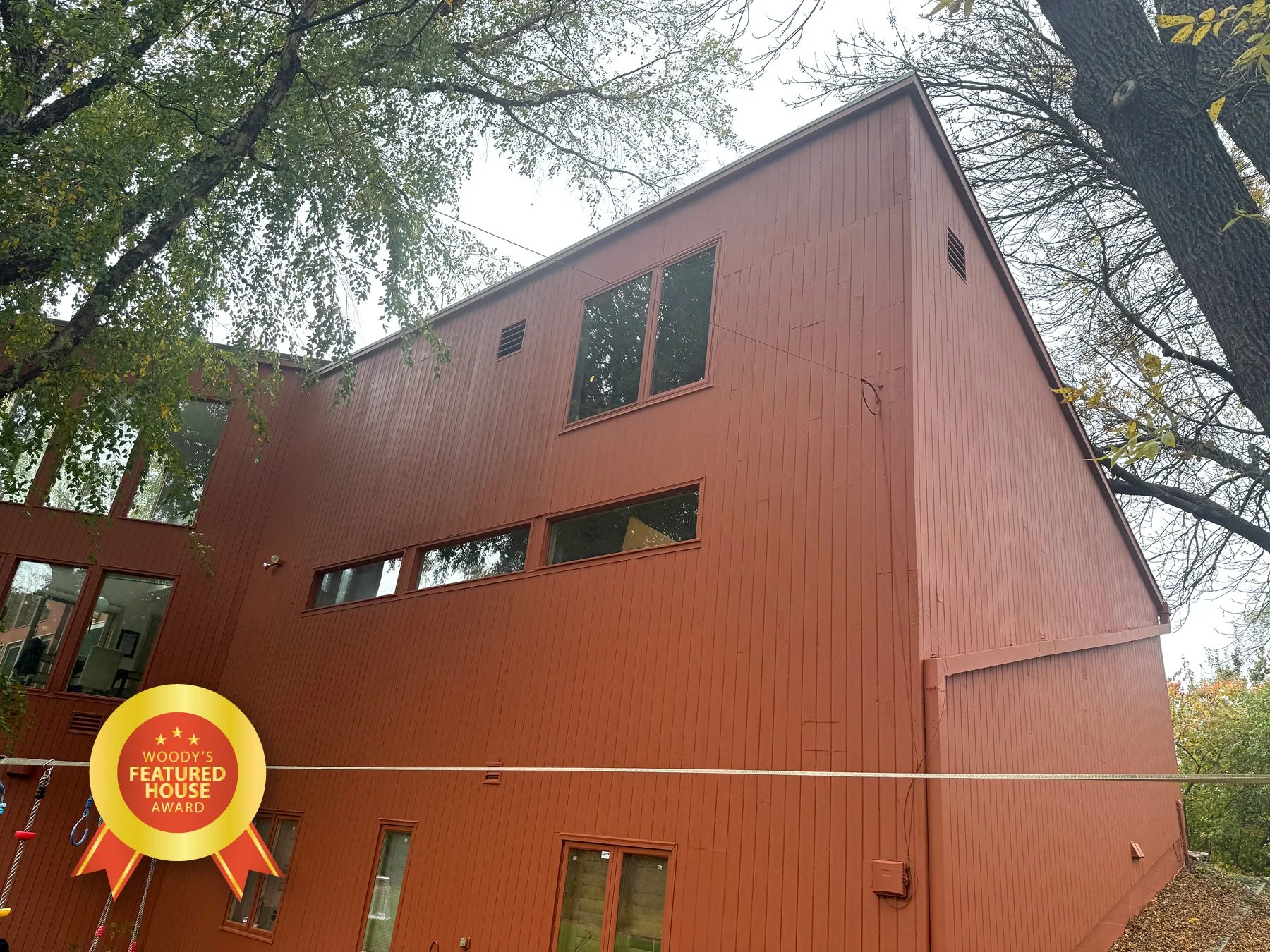 A three-story red wooden house with large windows, surrounded by trees with autumn foliage.