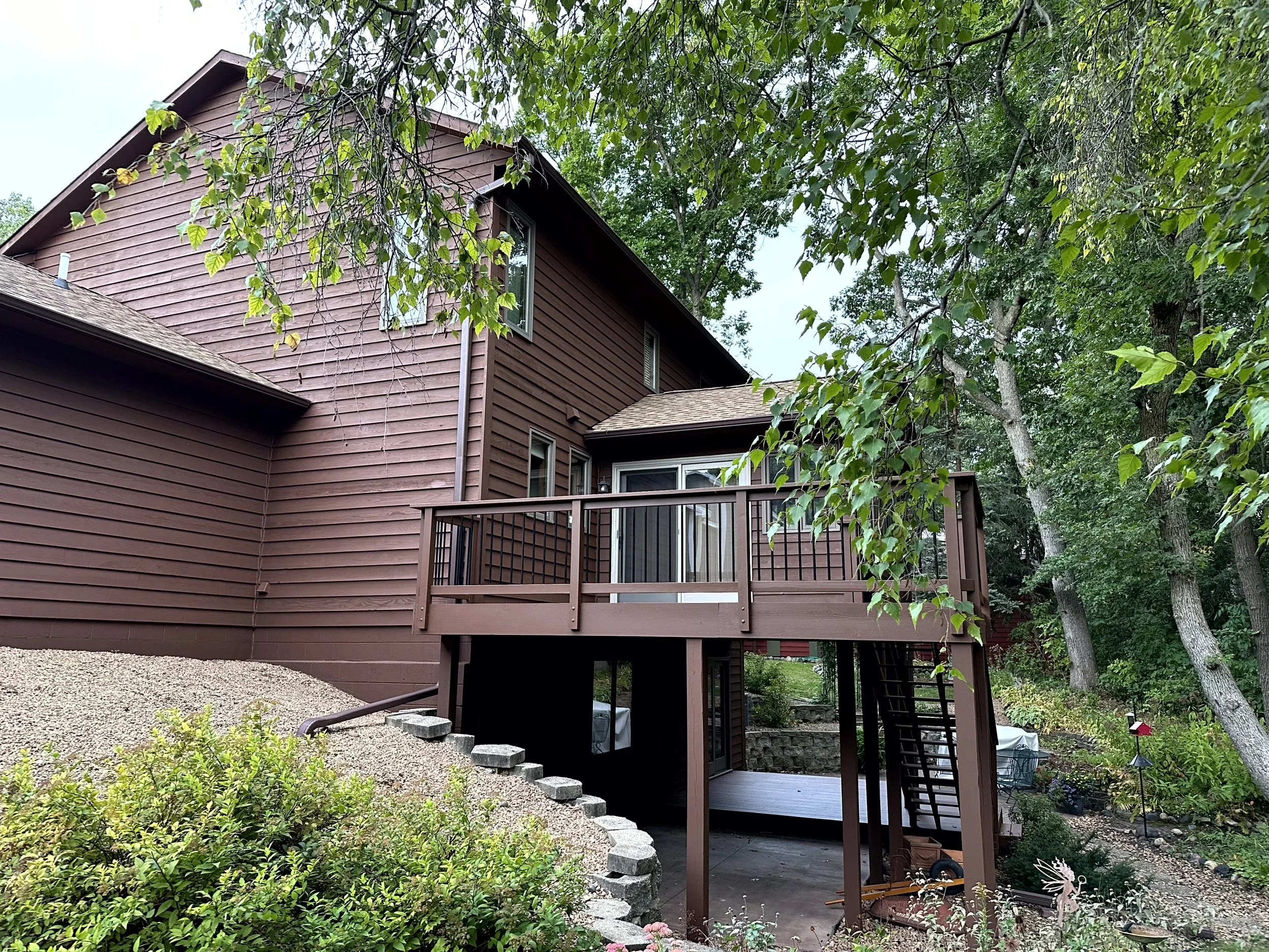 Backyard view of a two-story house with brown wooden siding, a small balcony, and ladder leading to the yard, surrounded by trees and plants.