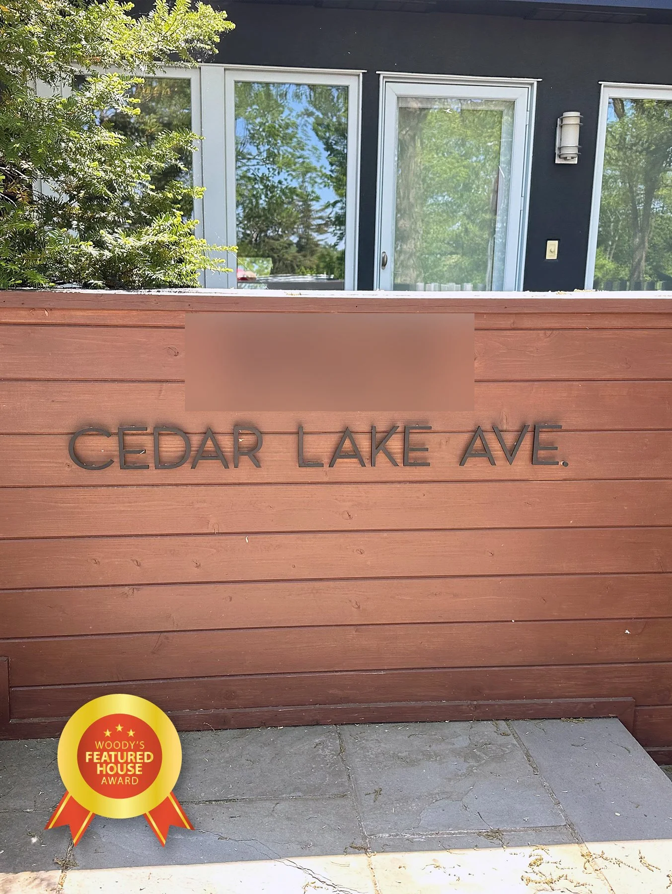 Wooden sign reads 'Cedar Lake Ave.' with a 'Woody's Featured House Award' ribbon sticker on the bottom left corner. In the background, there is a house with large windows and a glass sliding door, surrounded by greenery and trees.