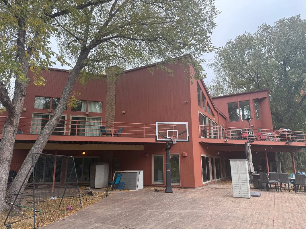 A multi-story red house with large windows and a spacious upper deck featuring railing, outdoor furniture, and a basketball hoop in the backyard. Trees surround the property.
