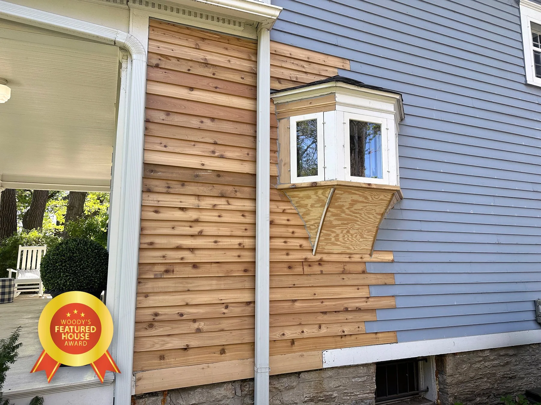 An exterior house wall with a bay window that is under construction, showing partially completed siding. The siding is being replaced from original gray to new natural wood. The house has blue siding on the right and white trim with a stone foundatio